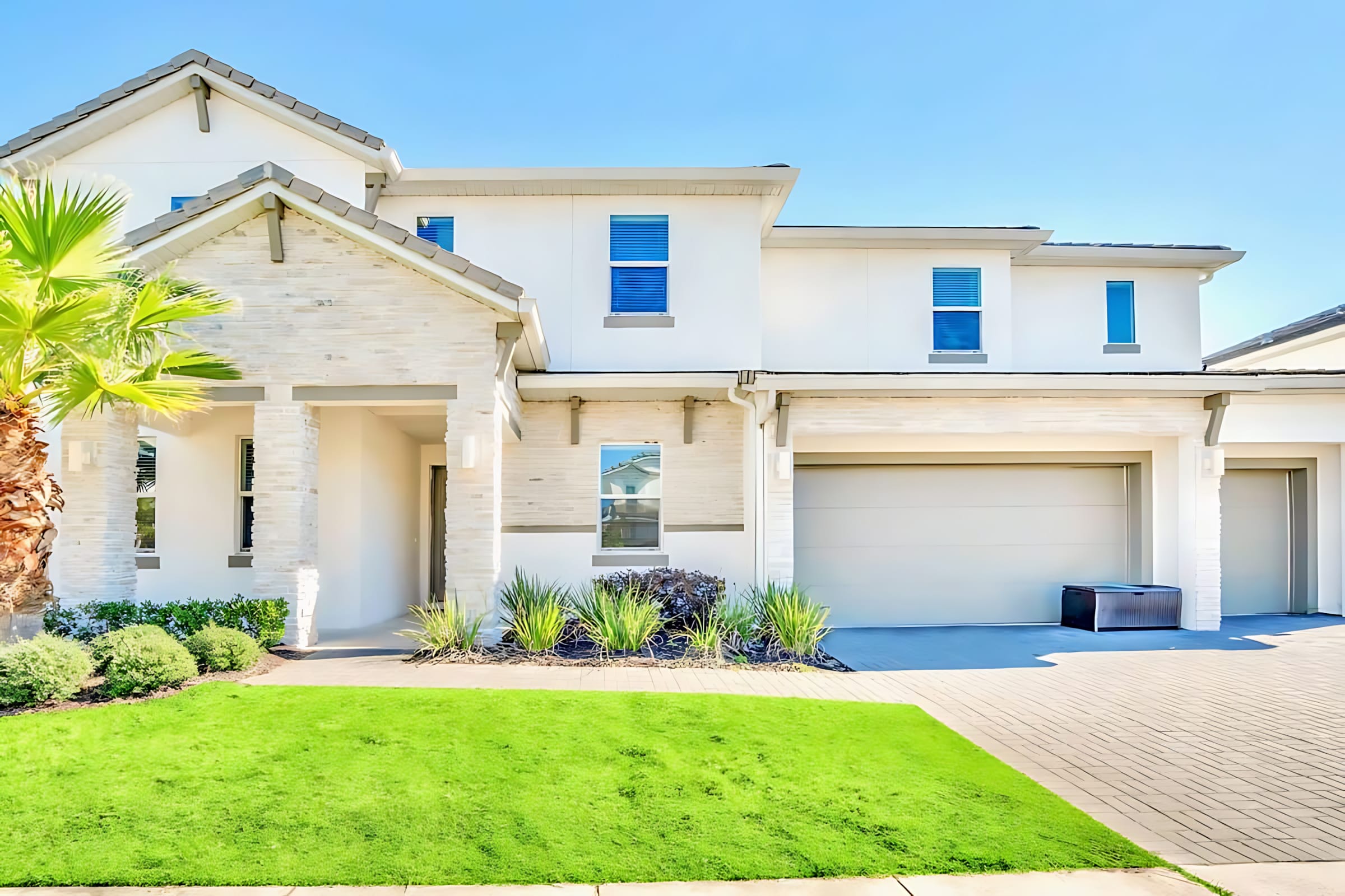 Modern two-story house exterior with white stucco and light stone accents, featuring a well-maintained green lawn and a paved driveway leading to a two-car garage. A palm tree adds a tropical touch to the landscaping.