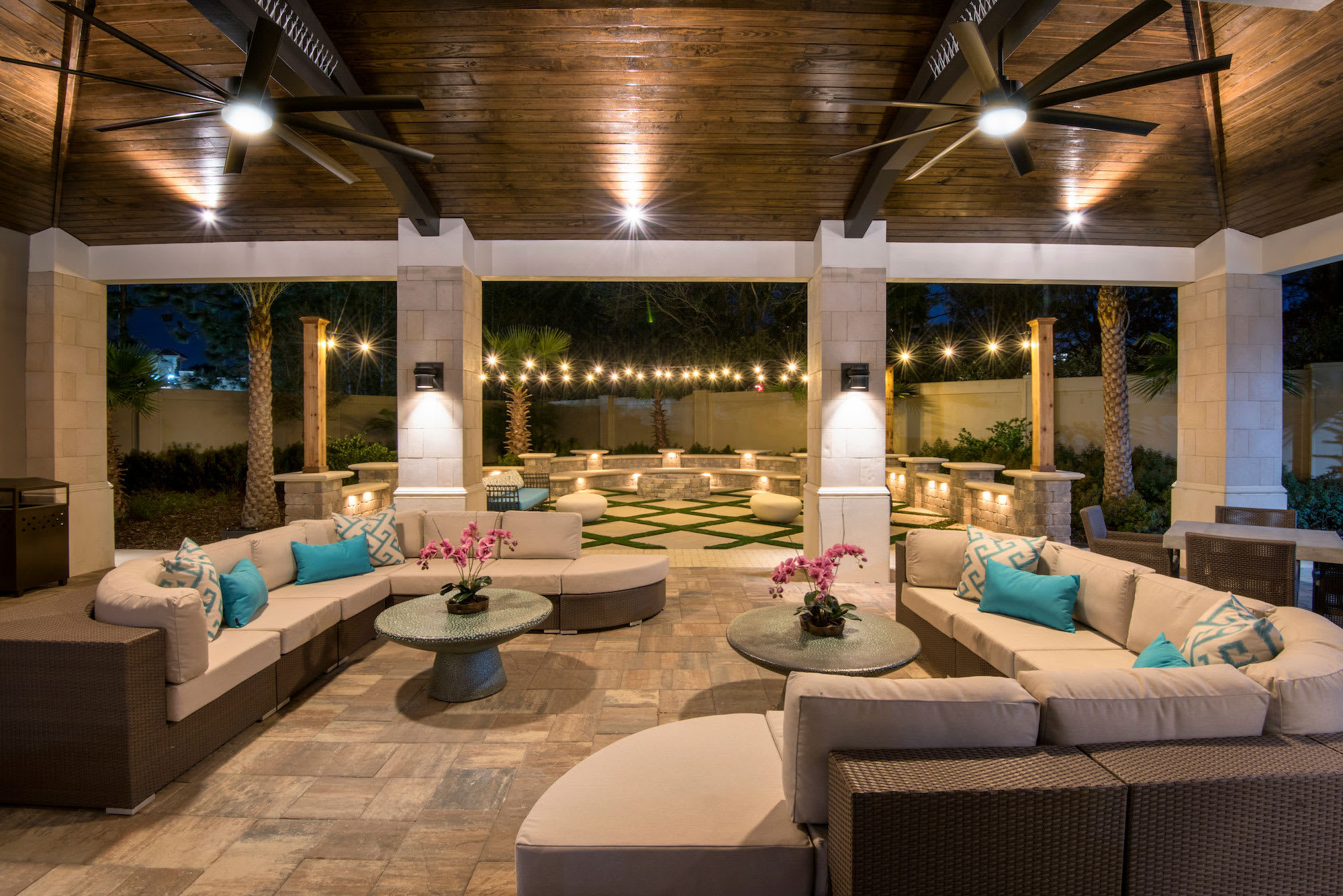 A spacious, covered outdoor patio lounge area featuring multiple comfortable wicker sofas with light cushions and blue accent pillows. Ceiling fans, string lights, and a fire pit area in the background create an inviting evening ambiance.