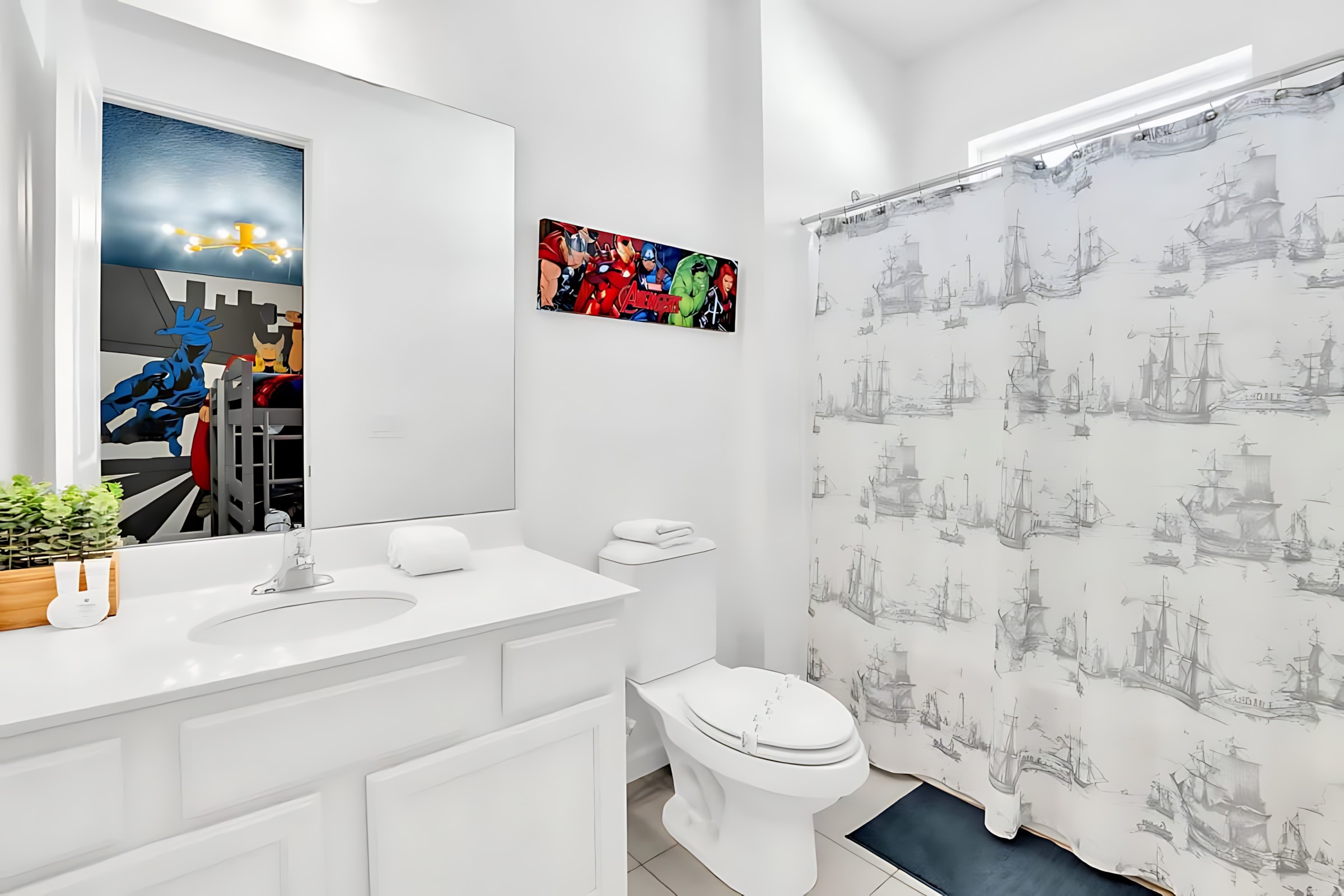 White modern bathroom with a single vanity, toilet, and a shower with a ship-themed curtain. A mirror reflects a colorful superhero-themed bedroom.
