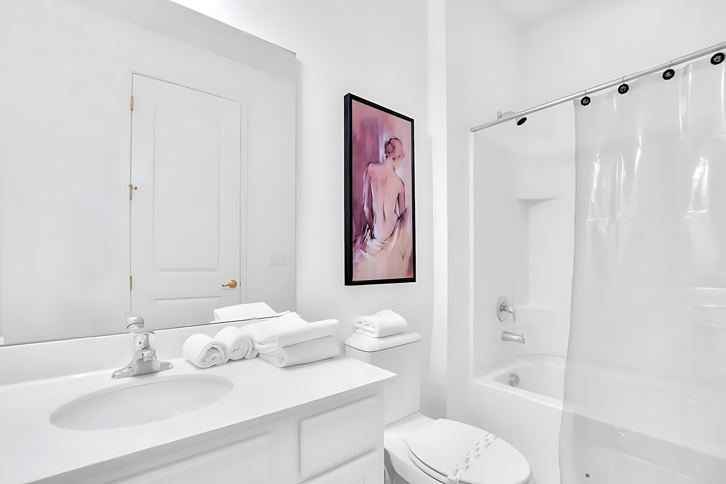 Minimalist white bathroom with a single vanity, toilet, and a shower/tub combination with a clear curtain. A framed art piece hangs above the toilet.