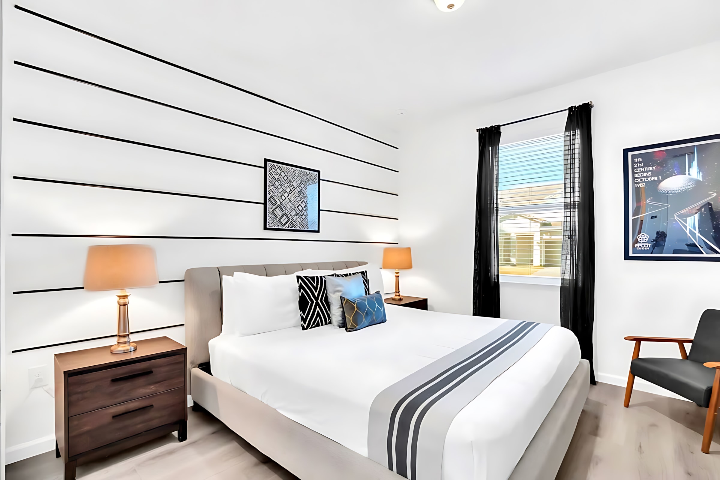 Modern bedroom with a queen-sized bed featuring a grey upholstered headboard, white bedding with black and grey accents, and black horizontal stripes on a white accent wall.