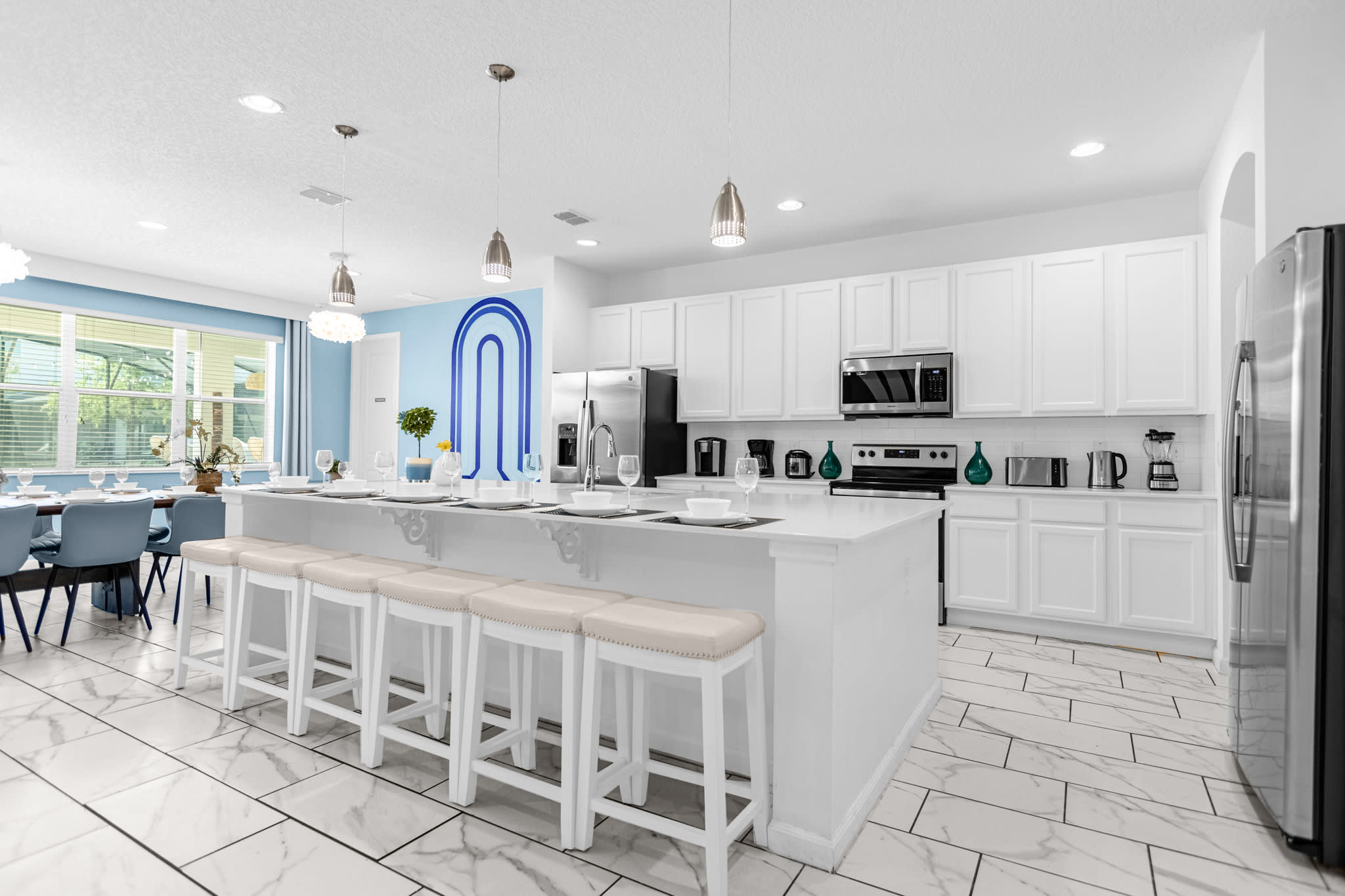 A modern white kitchen with sleek white cabinetry, white quartz countertops, stainless steel appliances, and a large island with six bar stools, extending into a dining area with a light blue accent wall.