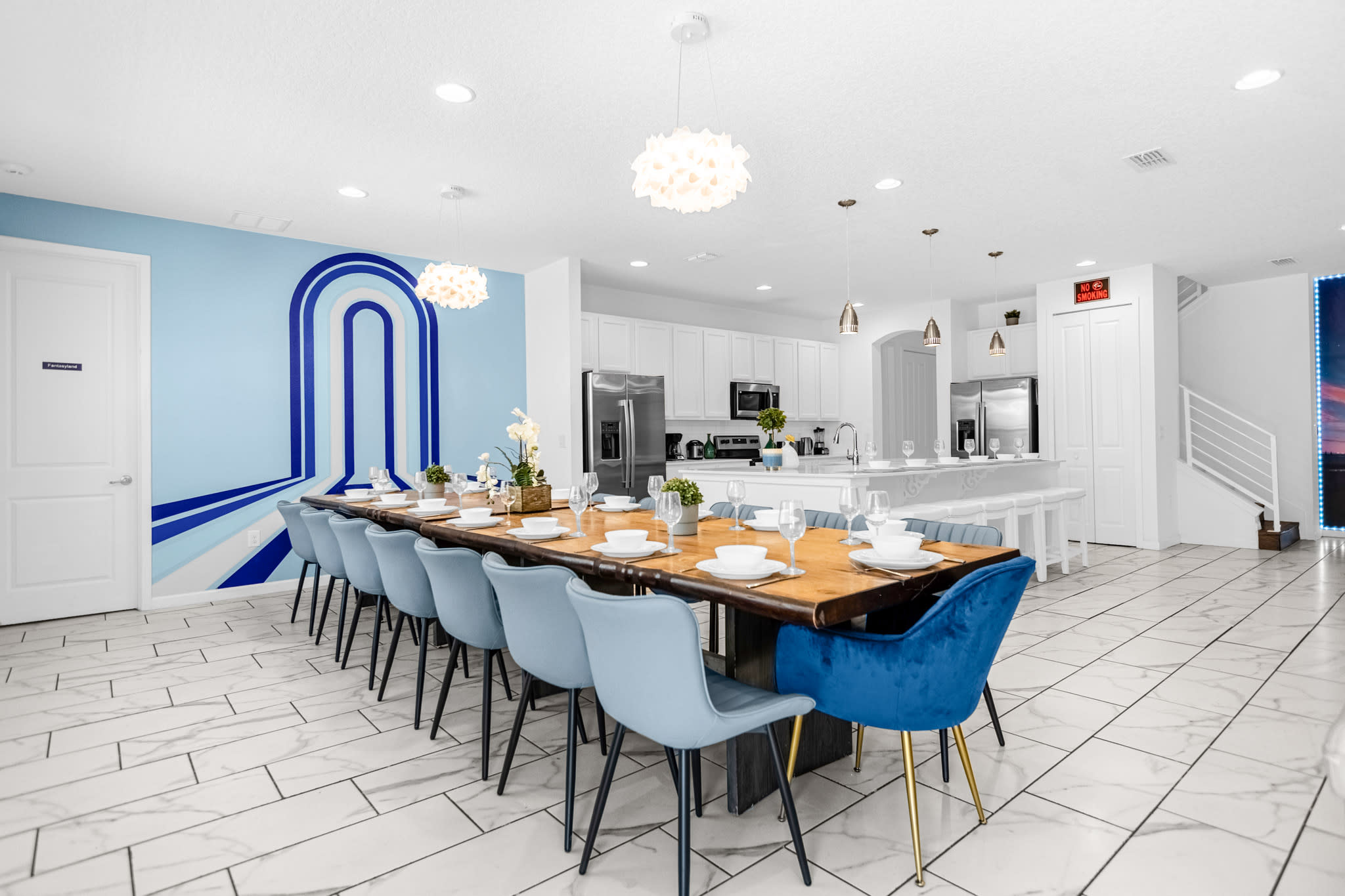 A spacious dining room featuring a long wooden dining table with seating for many, complemented by blue upholstered chairs, set against a light blue accent wall with a modern geometric design, and illuminated by contemporary pendant lights.
