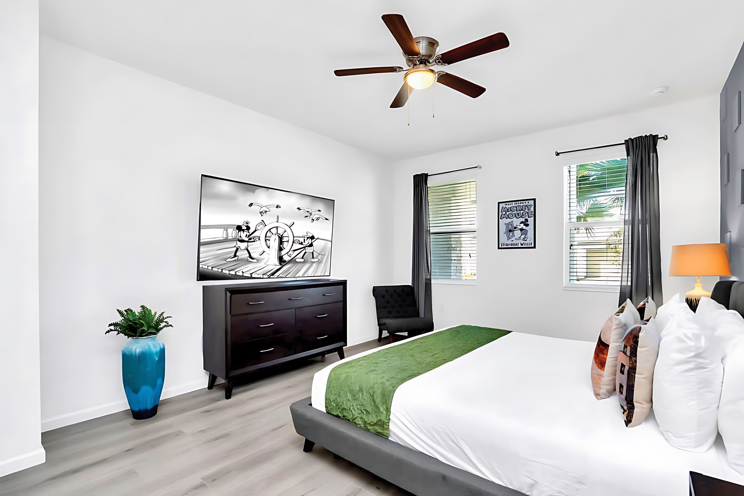 Bright and spacious bedroom with white walls, light wood-look flooring, a large wall-mounted TV above a dark dresser, and a bed with a green runner. A ceiling fan and an accent chair complete the room.