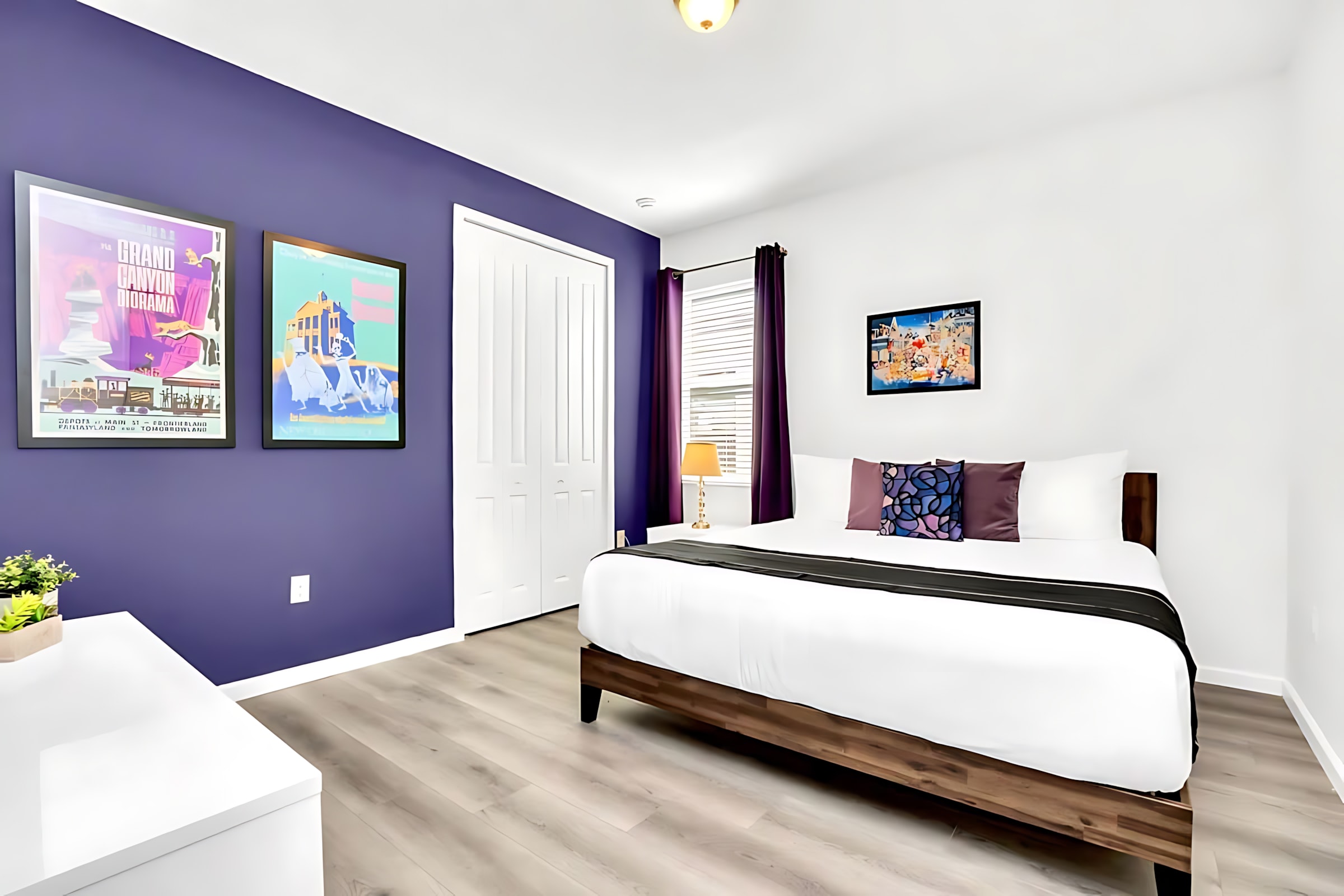 Modern bedroom with a queen-sized bed, dark wood frame, and a striking purple accent wall, complemented by two framed art prints and a white bi-fold closet.