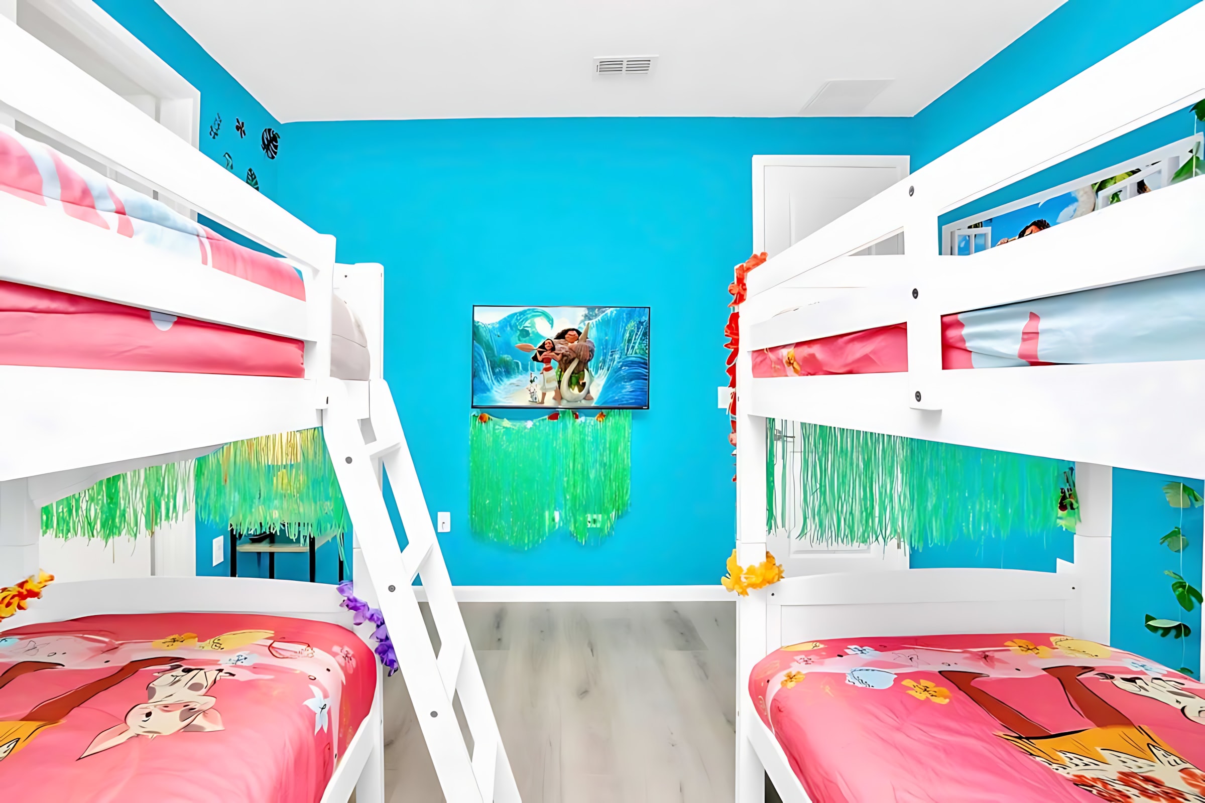 Close-up view of the Moana-themed bedroom featuring two white bunk beds with pink bedding, bright blue walls, and a wall-mounted TV displaying a Moana scene.