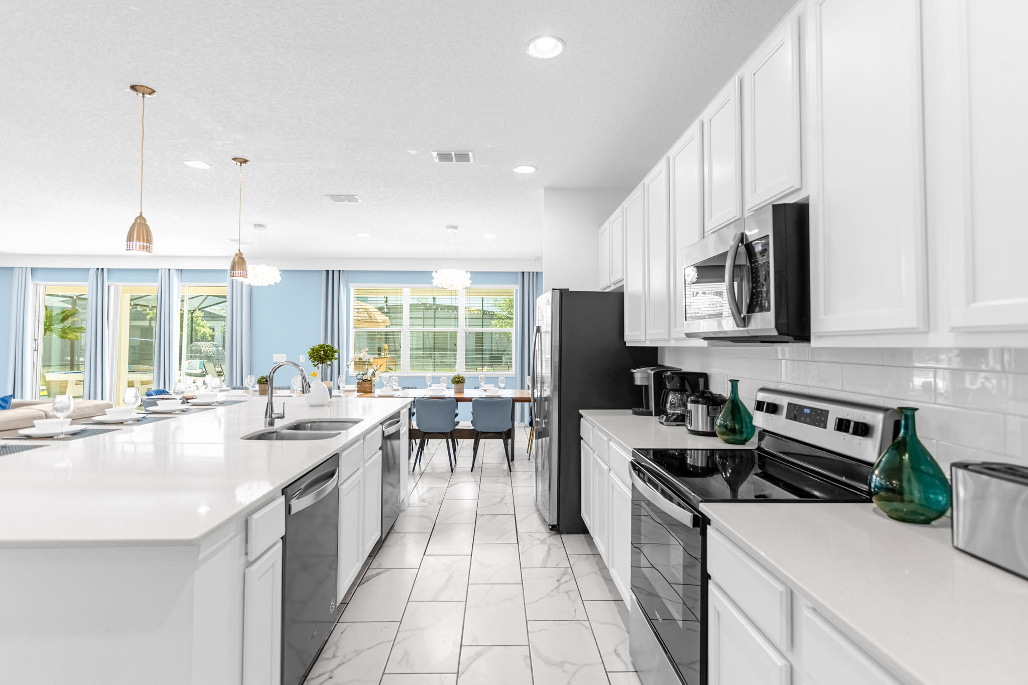 A detailed view of the modern white kitchen showcasing white shaker cabinets, white countertops, and a suite of stainless steel appliances including a refrigerator, microwave, oven, and dishwasher, with various small kitchen appliances on the counter.
