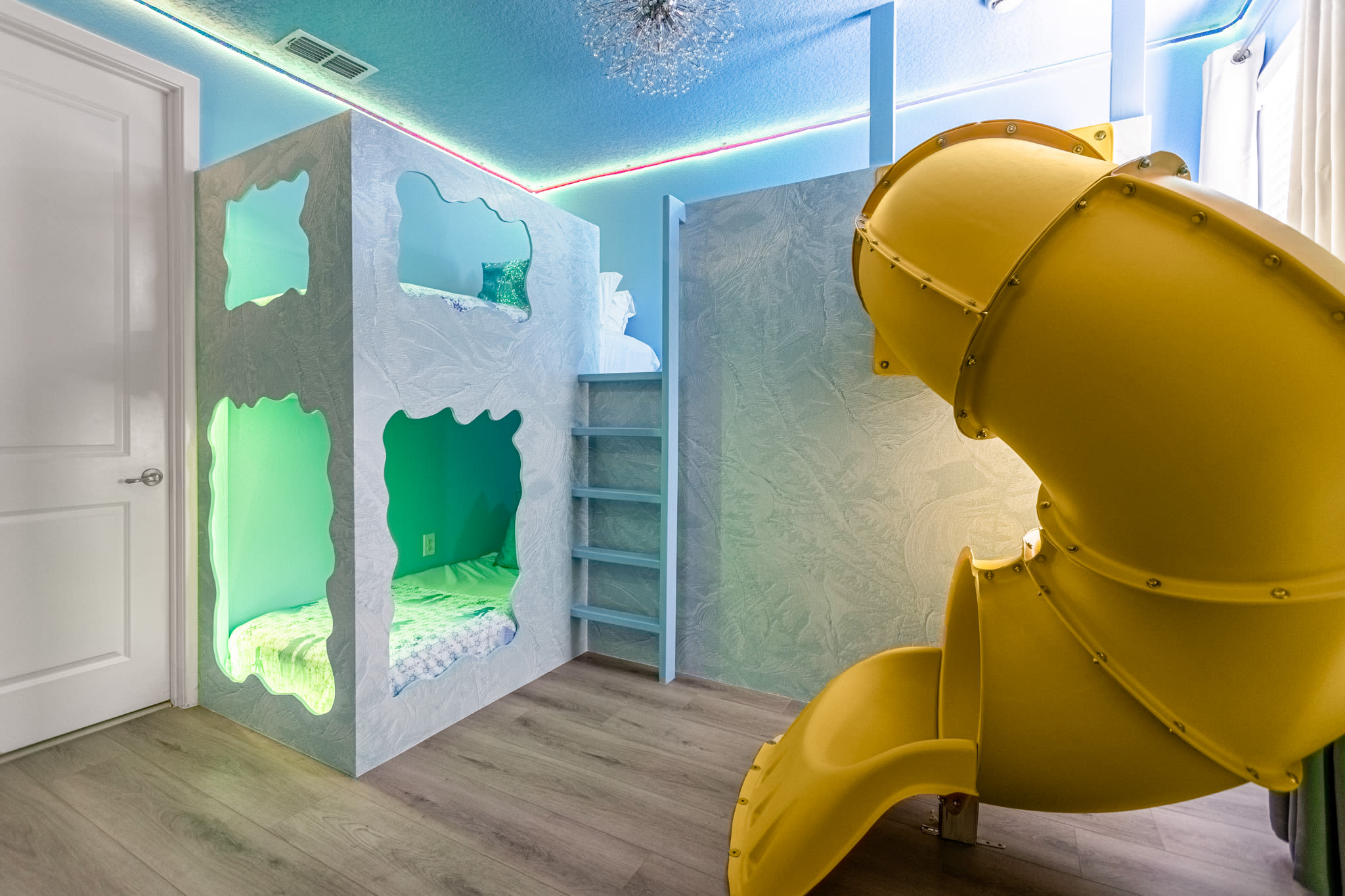 Detailed view of the 'Frozen' themed bedroom, showcasing the unique custom bunk beds with illuminated sleeping nooks and the large yellow slide. The room features light blue walls and wood-look flooring.