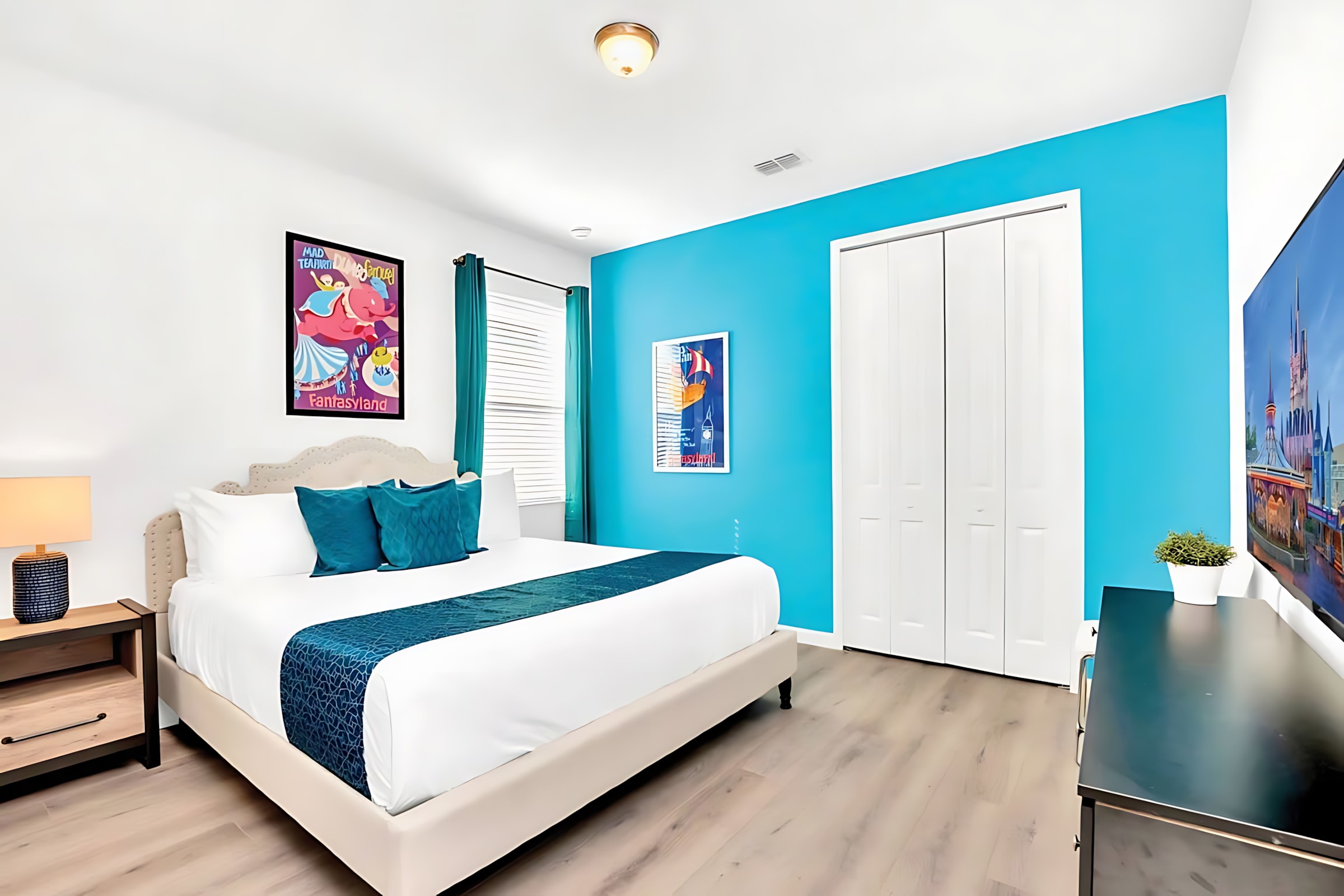 Bright bedroom with a queen-sized bed, white bedding, and a vibrant blue accent wall, featuring a large flat-screen TV and a white bi-fold closet.