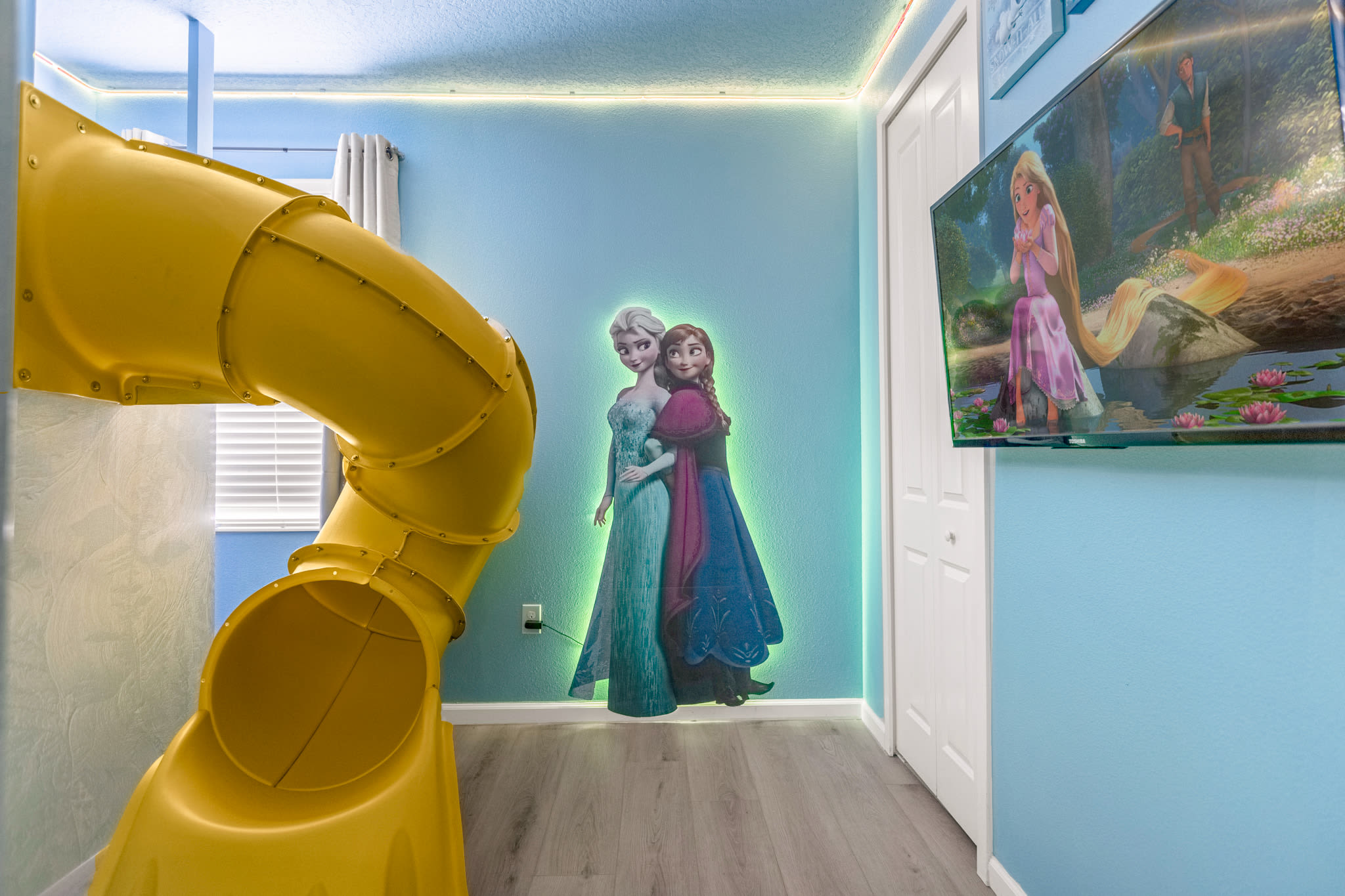 Corner view of the 'Frozen' themed bedroom, highlighting the character cutouts of Elsa and Anna, a wall-mounted TV, and the base of the yellow slide. The walls are light blue.