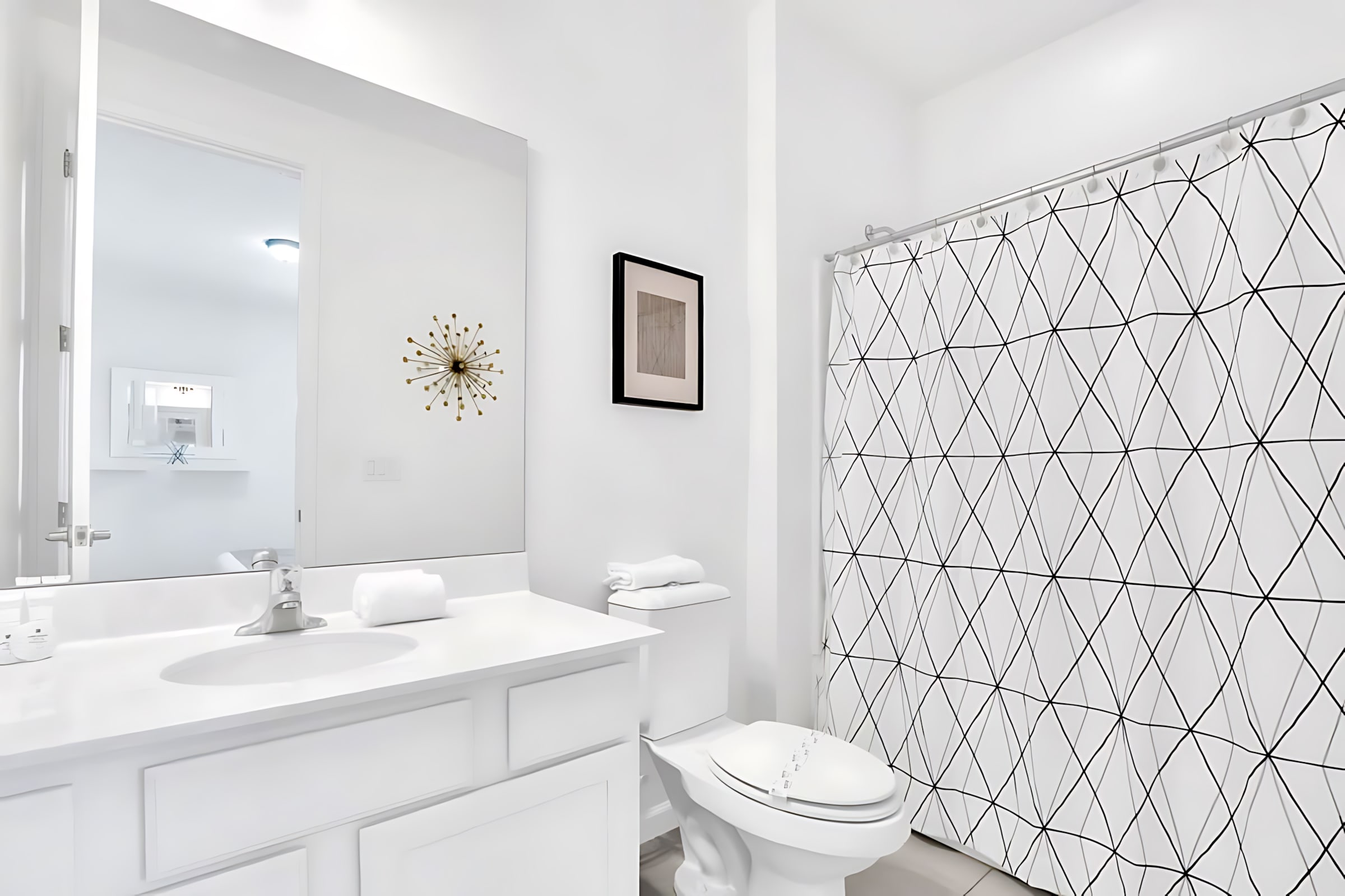 A clean, white bathroom featuring a vanity with a sink and a large mirror. A toilet is next to a shower enclosed by a modern geometric pattern curtain, with a small framed artwork and a decorative wall hanging adding visual interest.