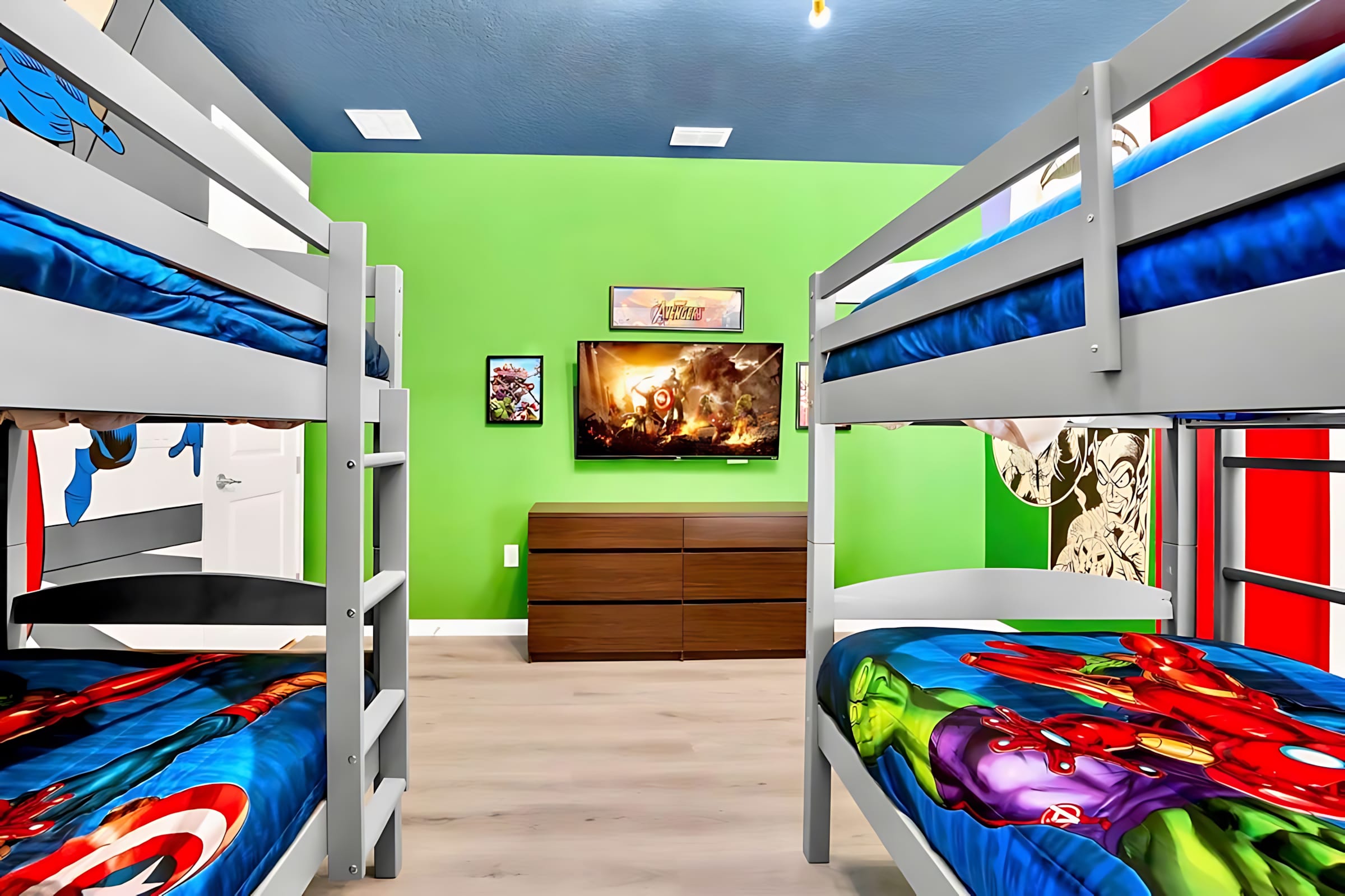 View of the superhero-themed bedroom showing two bunk beds with superhero bedding, a bright green accent wall, a wall-mounted TV, and a wooden dresser. The room has light wood-look flooring.