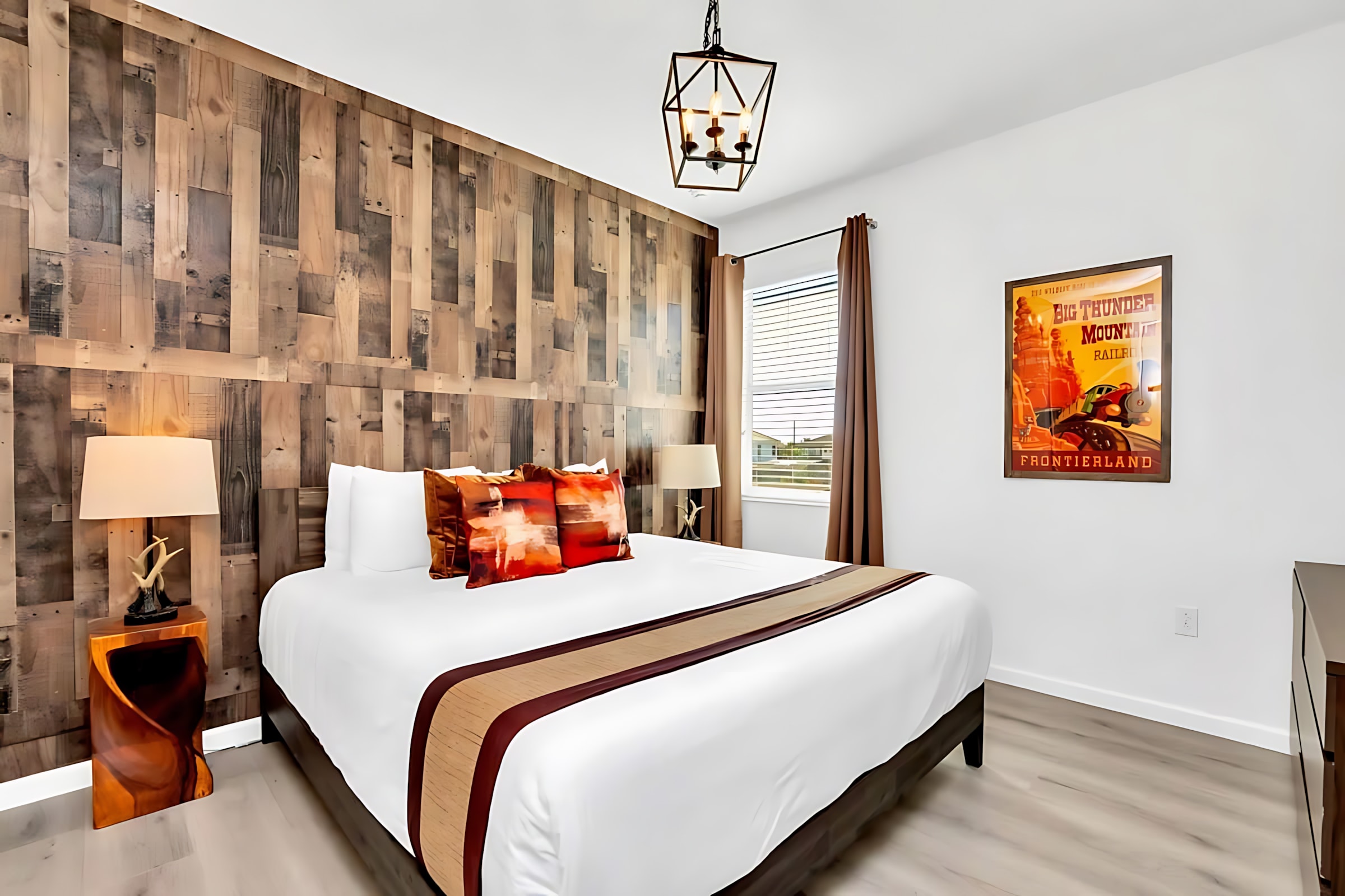 A modern bedroom featuring a king-sized bed with a white and brown runner, flanked by nightstands with unique lamps. A striking wooden plank accent wall serves as the headboard, complemented by a framed "Big Thunder Mountain Railroad" poster on the white wall.