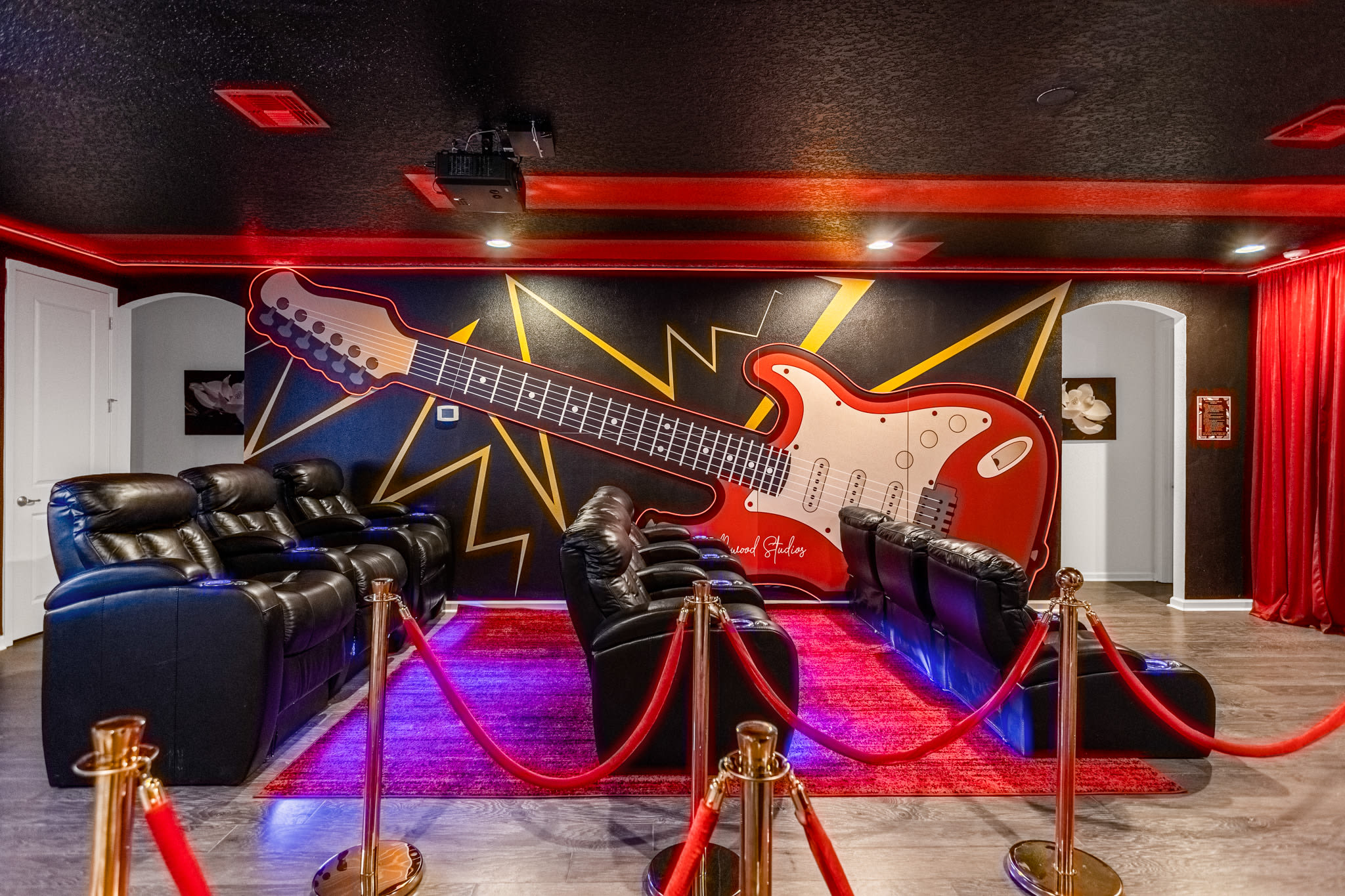 The media room features a large red and black guitar mural on the wall, plush black recliner seating arranged in rows, and red velvet ropes with stanchions leading to the viewing area, all under a dark ceiling with red accent lighting.