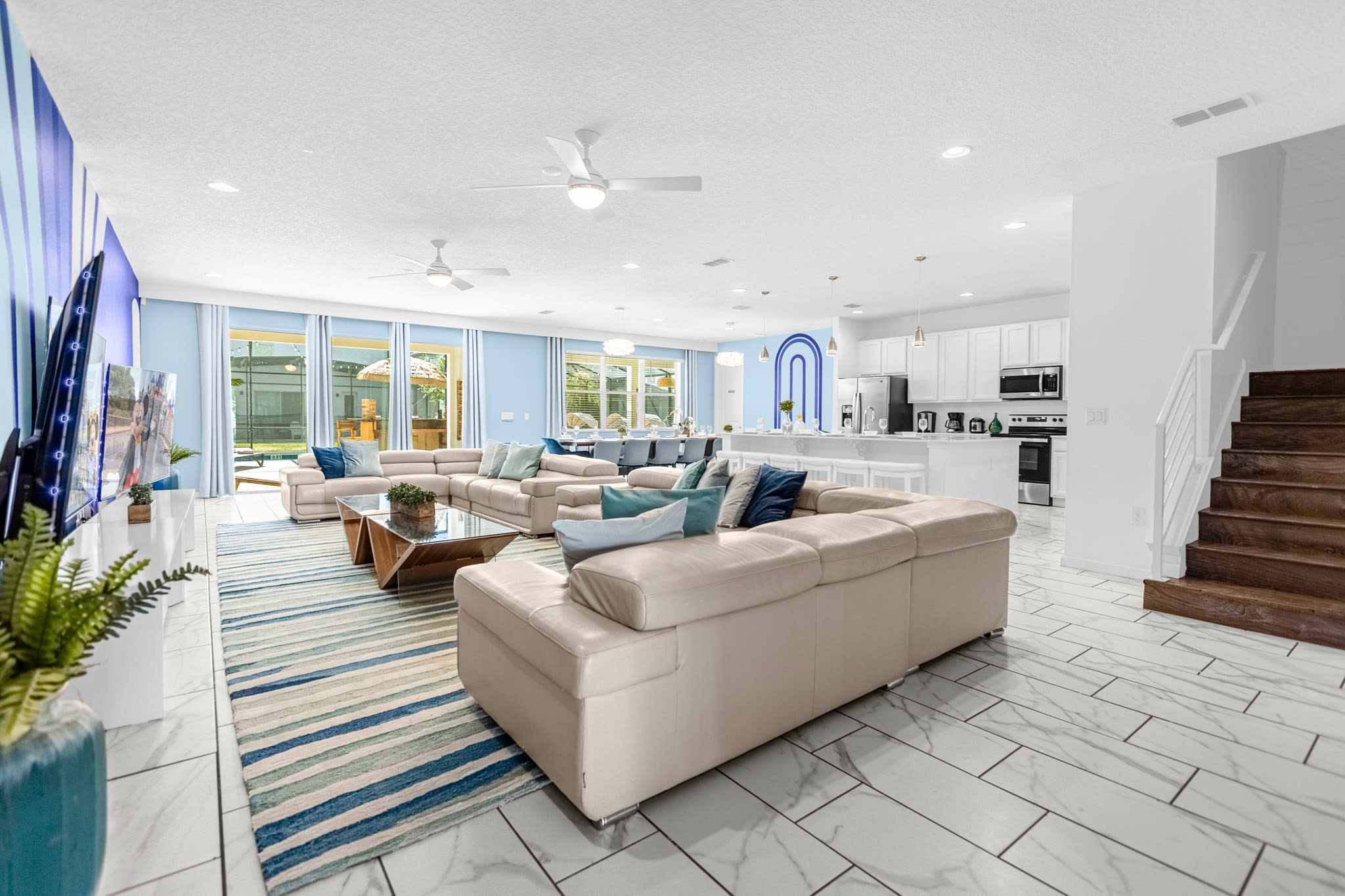 A bright and spacious open-concept living area with light blue walls, white tiled floors, multiple cream-colored sofas, and a blue and white striped rug, leading into a modern white kitchen and dining area with large windows overlooking the outdoors.