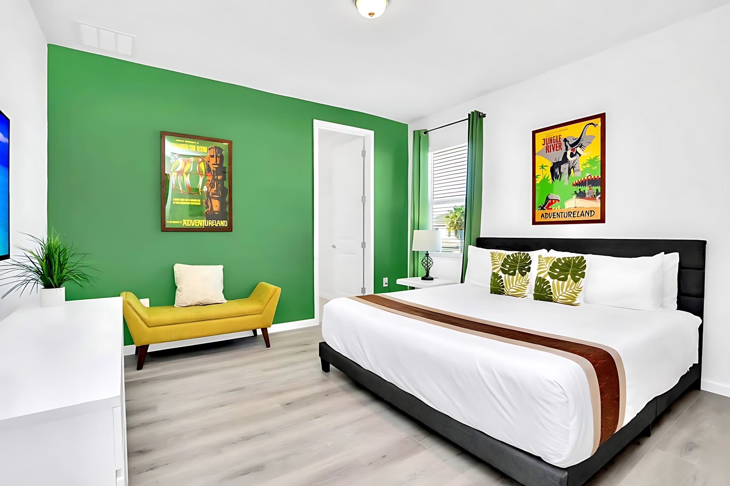 Spacious bedroom with a king-sized bed, white bedding, and a bold green accent wall, featuring a yellow upholstered bench and a large flat-screen TV.