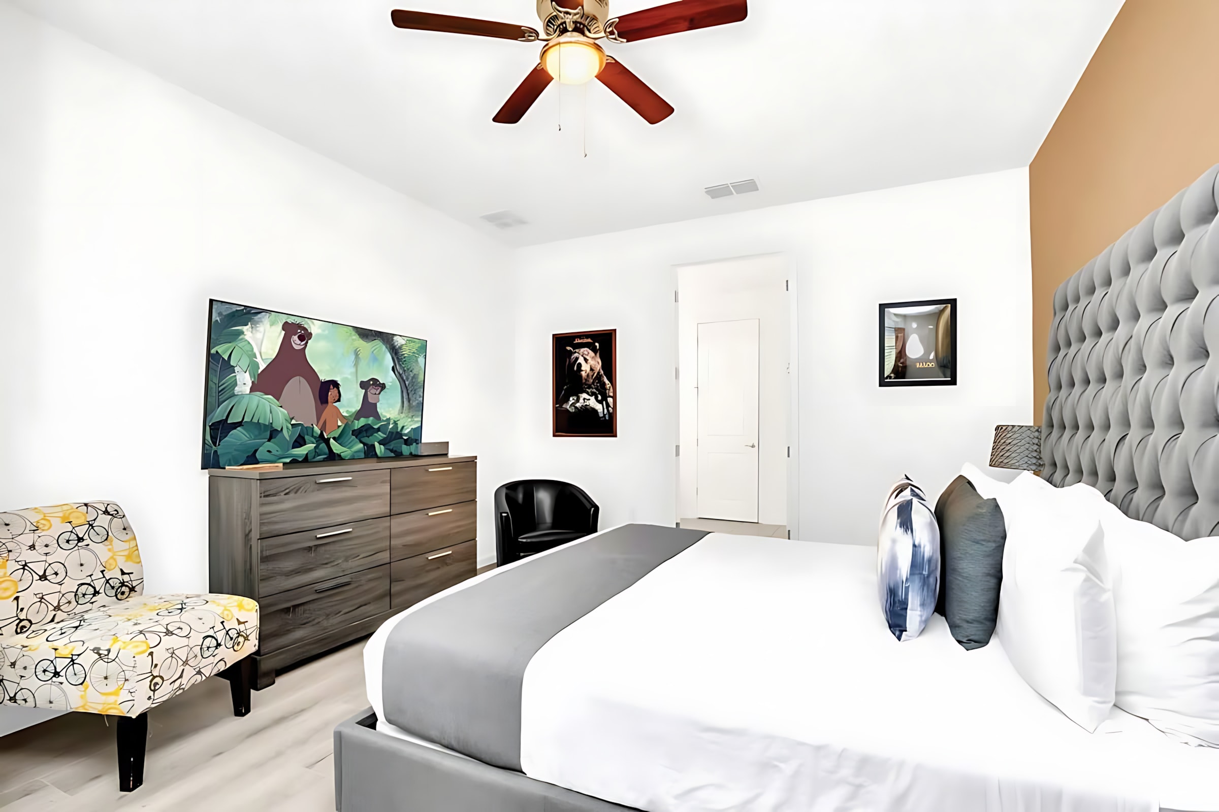 Another perspective of the bedroom, highlighting the king-size bed with a gray tufted headboard, a large dresser with a flat-screen TV displaying a cartoon, a patterned armchair, and a ceiling fan, with artwork adorning the white walls.
