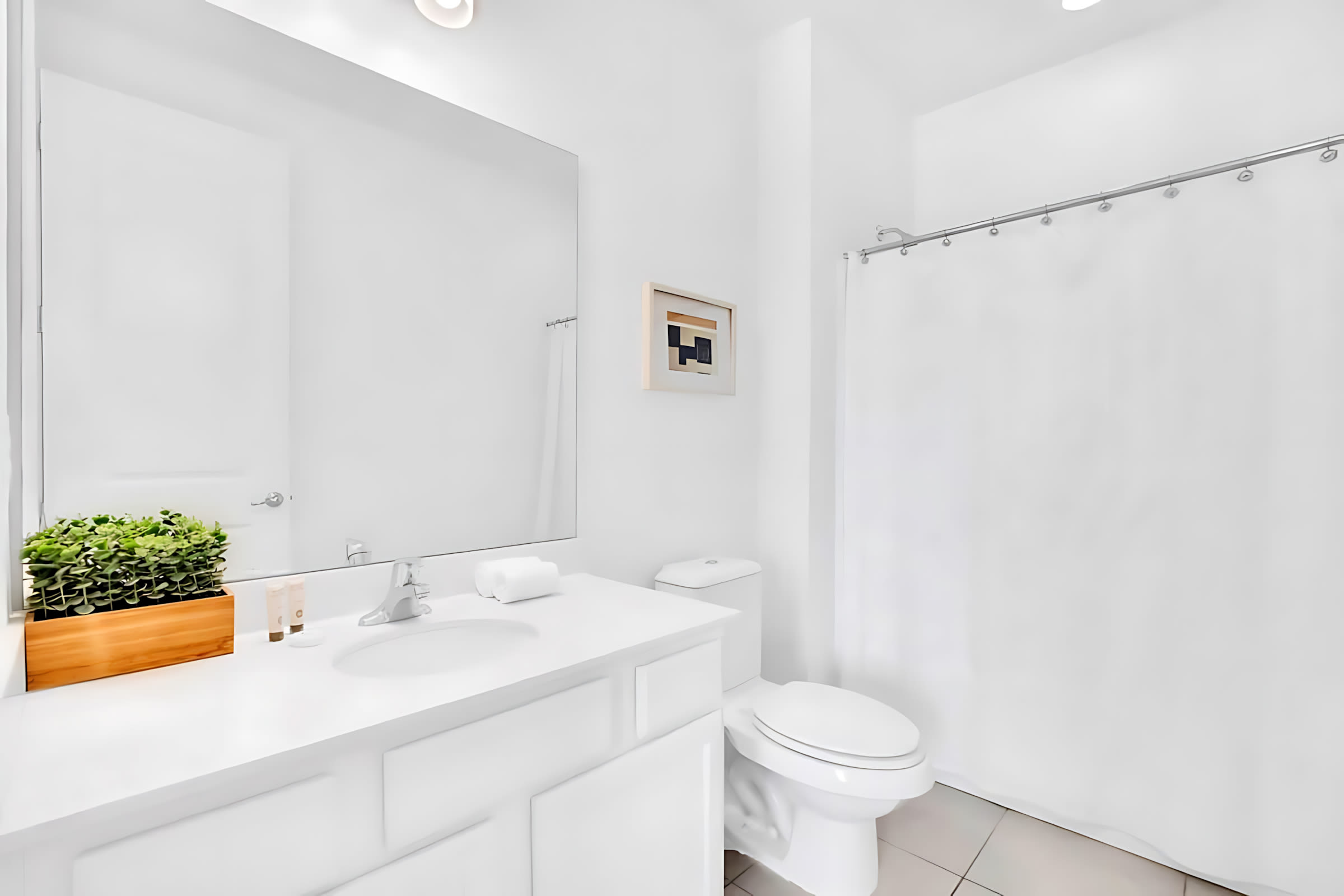 A bright, all-white bathroom with a modern vanity, sink, and large mirror. A toilet is positioned next to a shower with a simple white curtain, and a small framed artwork adds a touch of decor.