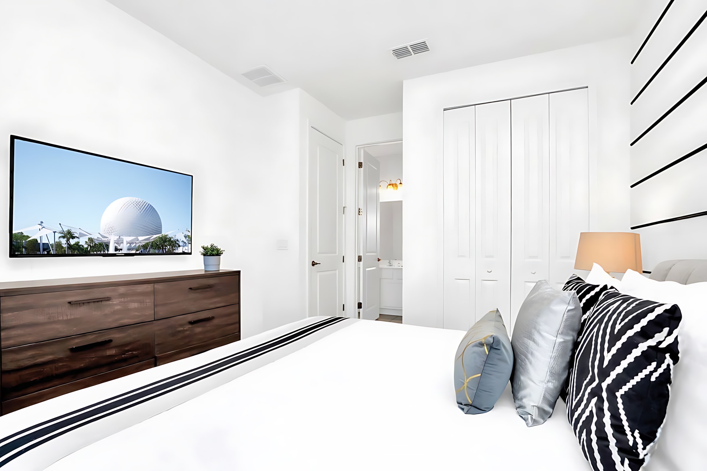 View of the modern bedroom showing a wall-mounted TV above a dark wood dresser, a queen-sized bed with decorative pillows, and a doorway leading to a white bathroom.