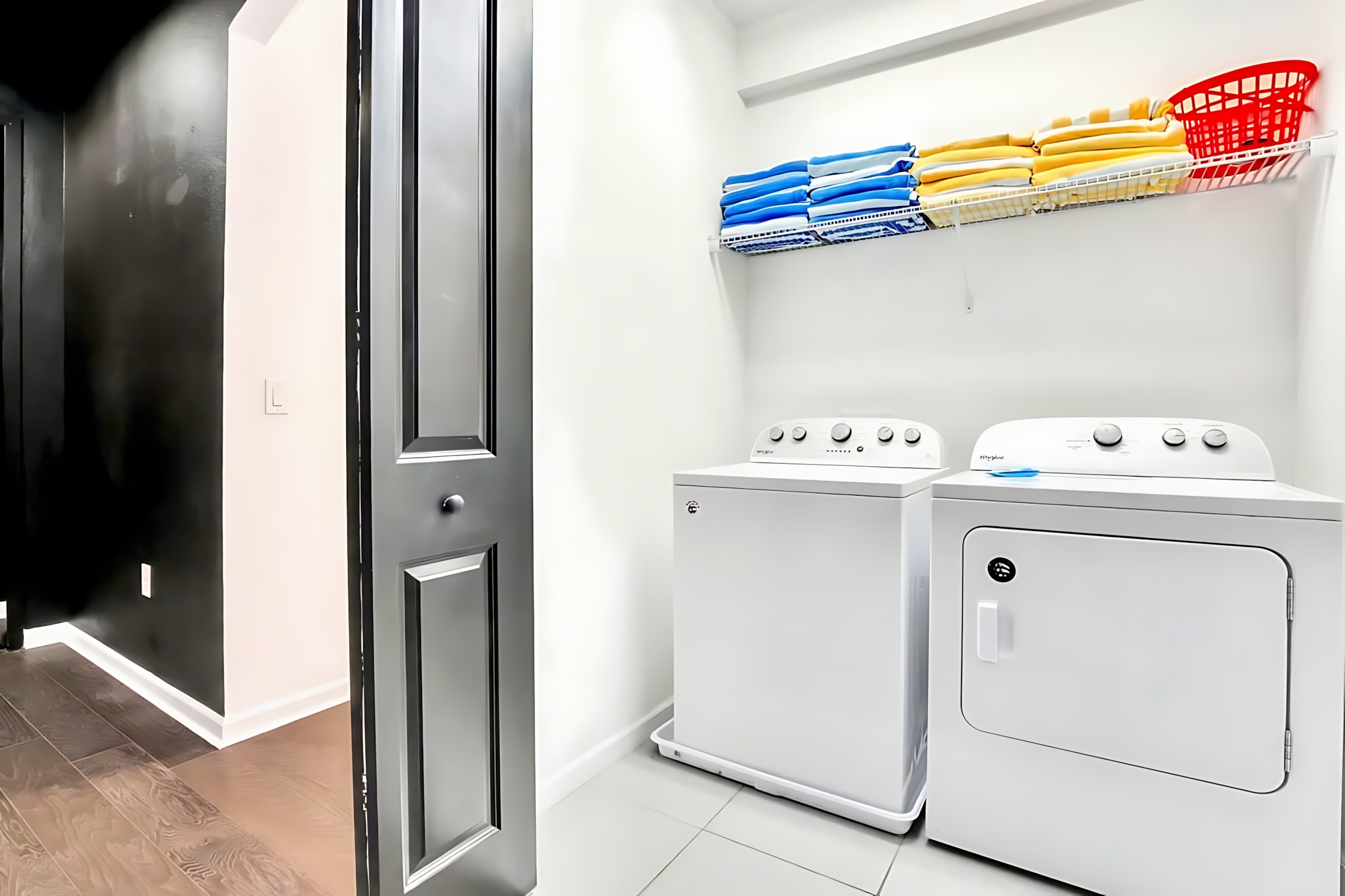 A functional laundry room equipped with a white washer and dryer. A wire shelf above holds neatly folded towels and a red laundry basket, with a dark door visible on the left.