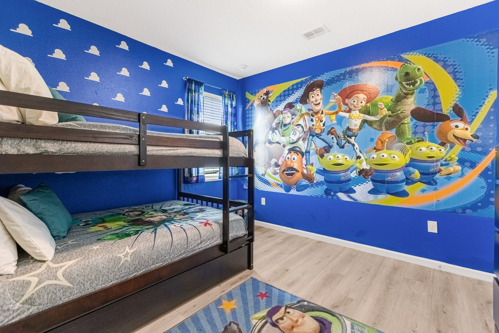 Wide view of the Toy Story-themed bedroom showcasing a large wall mural of characters, a dark wood bunk bed with themed bedding, and a blue wall with cloud patterns.