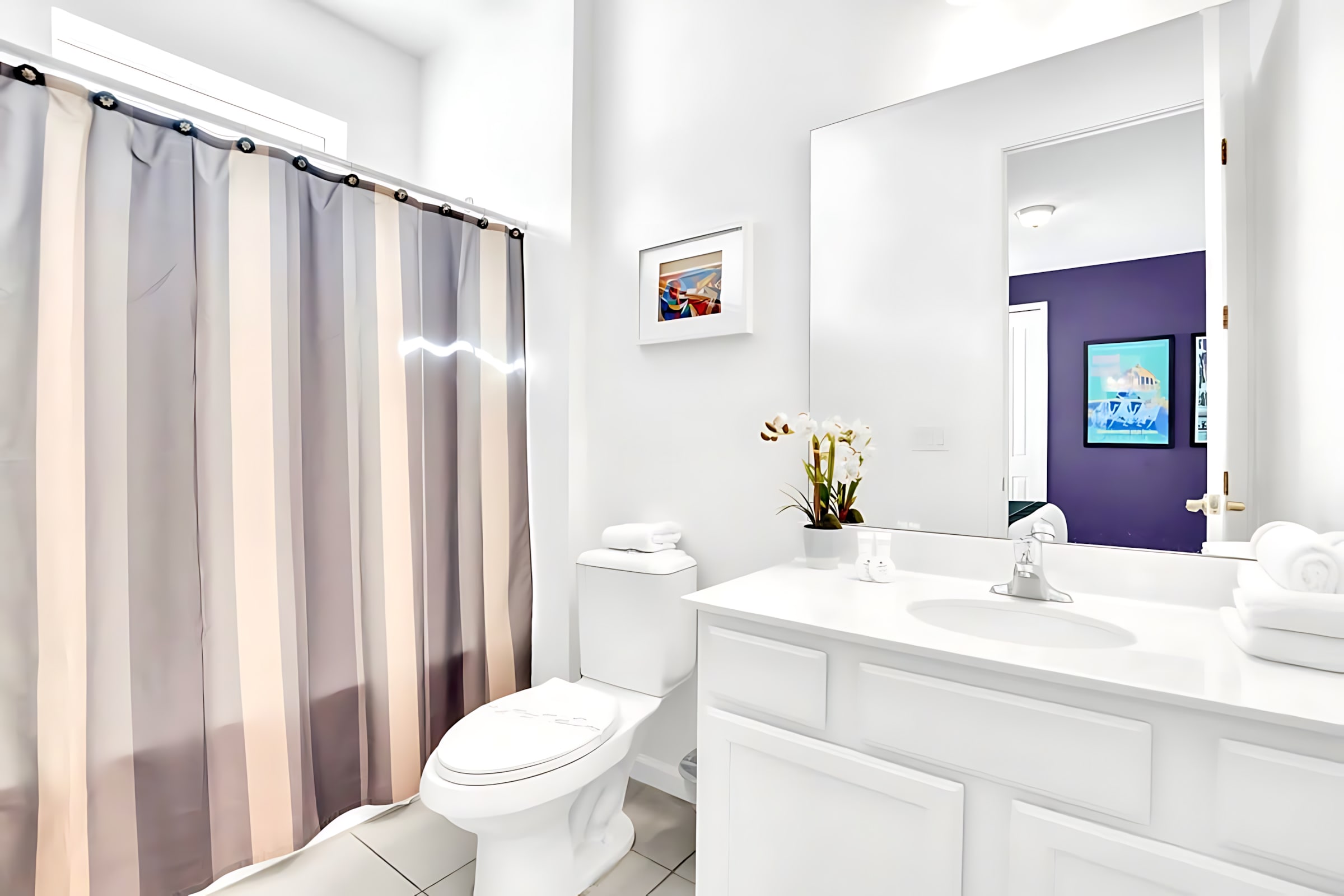 Bright white bathroom featuring a single vanity with a large mirror, a toilet, and a shower/tub combo enclosed by a grey and white striped shower curtain.