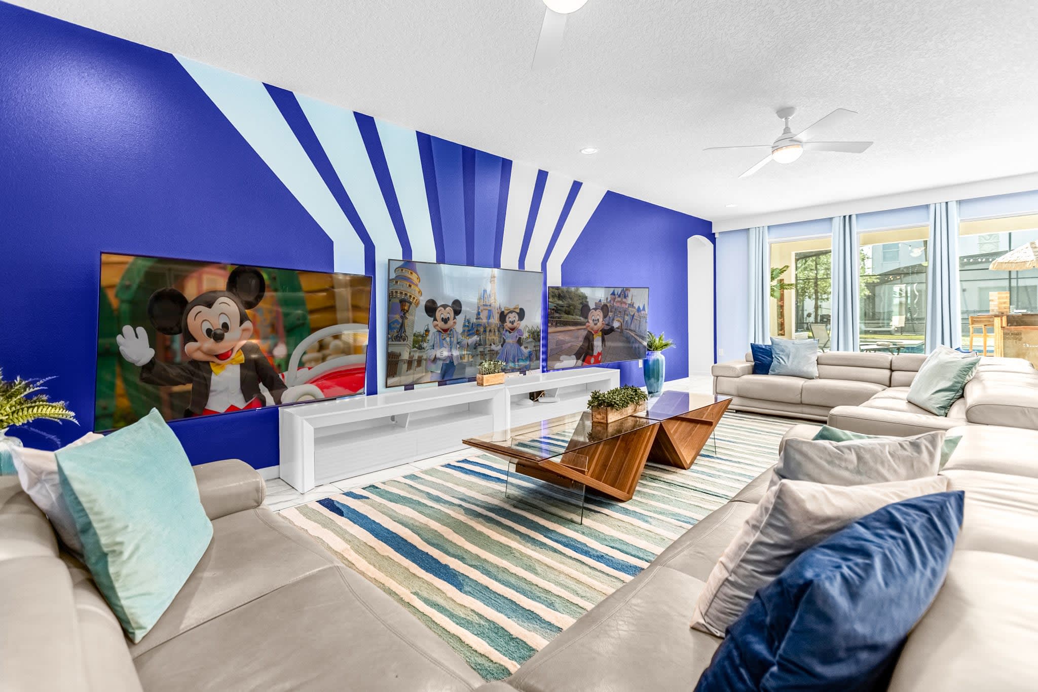 The living room features a striking blue accent wall with white geometric stripes, three large flat-screen TVs displaying Disney characters, and light gray sectional sofas arranged around a unique wooden and glass coffee table on a blue and white striped rug.