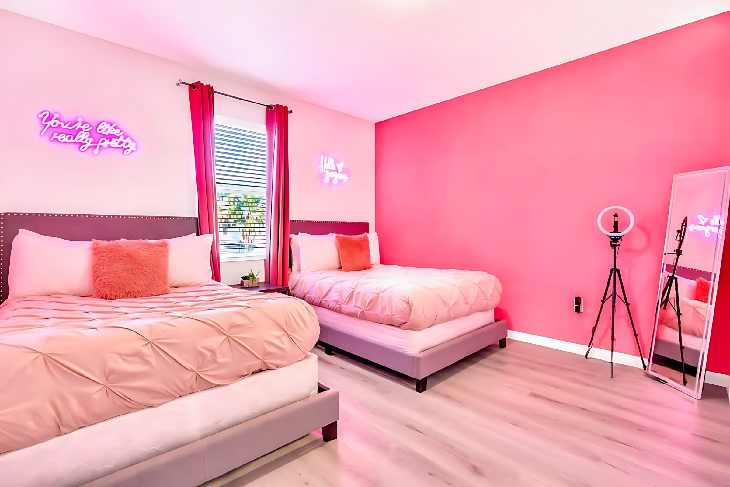 Vibrant pink-themed bedroom with two beds, light wood-look flooring, and playful neon wall signs. The room also features a ring light on a tripod and a full-length mirror.
