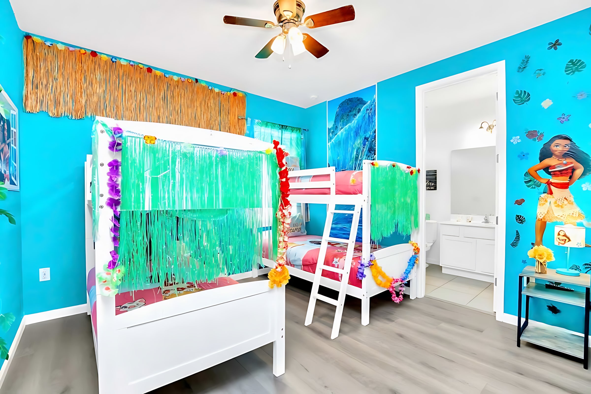 Vibrant Moana-themed bedroom with bright blue walls, two white bunk beds decorated with green and yellow garlands, and a ceiling fan. A doorway leads to a white bathroom.