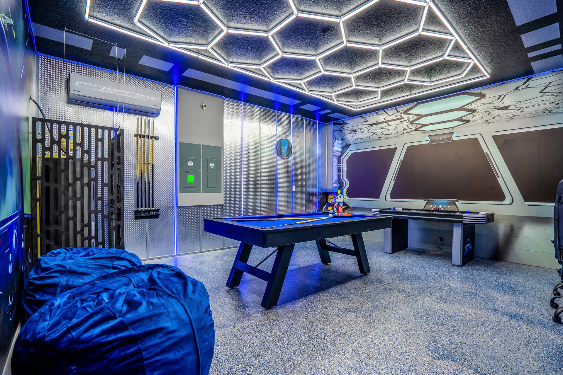 A wide shot of the sci-fi themed game room, showcasing a blue-felt pool table, a wall-mounted pool cue rack, and two large blue bean bag chairs, with hexagonal ceiling lights and metallic wall panels.
