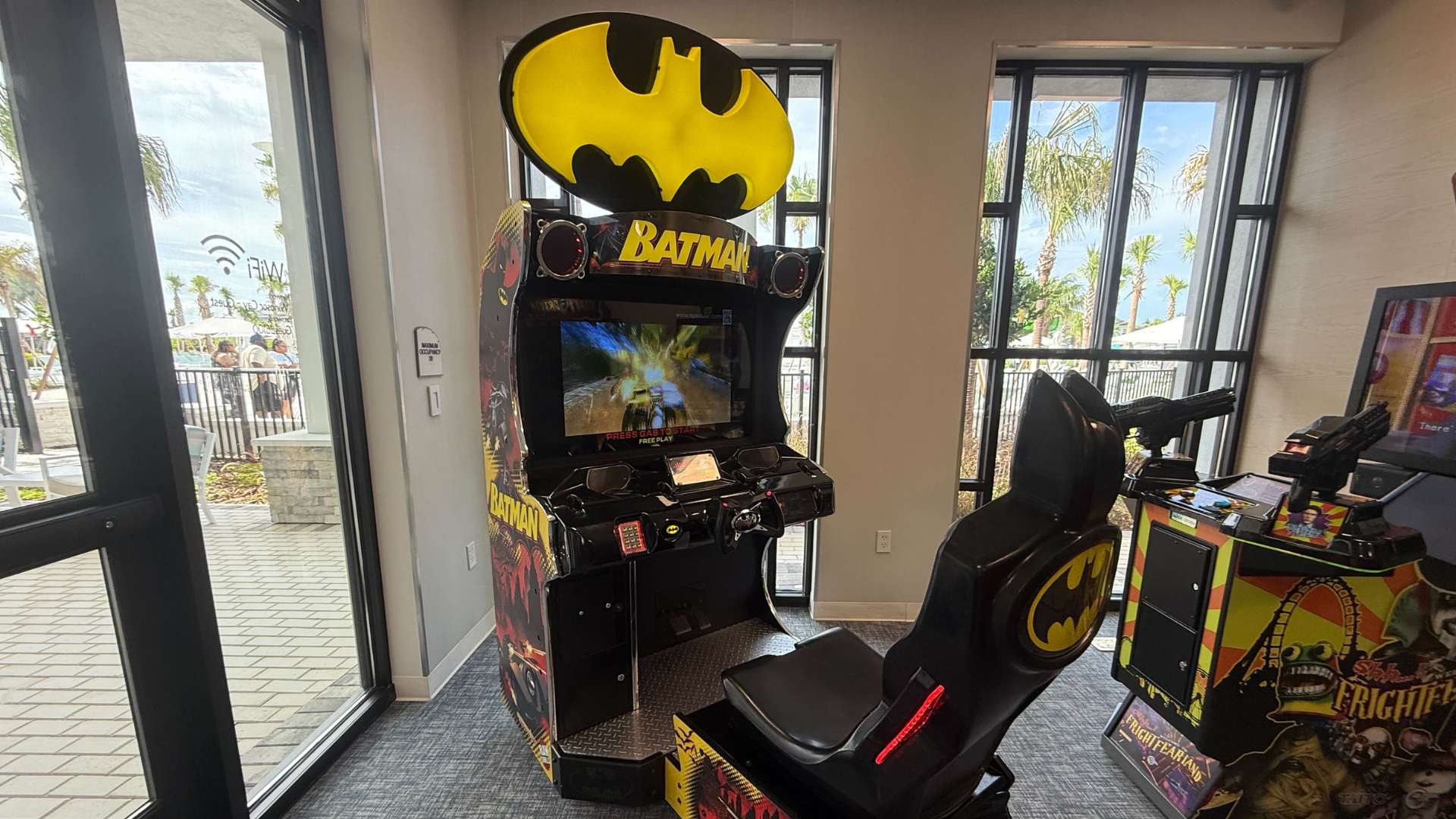 A close-up view highlights a Batman arcade racing game with a prominent yellow Bat-Signal marquee, featuring a black racing seat and controls. The game is positioned near large windows overlooking an outdoor pool area, with a gray patterned carpet and light wood-paneled walls.