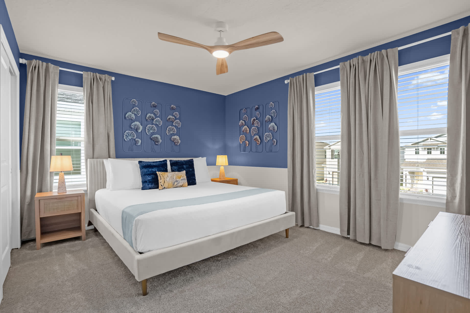 Vibrant bedroom with a king-sized bed, a striking blue accent wall with decorative metal wall art, two wooden nightstands with lamps, and large windows with gray curtains.