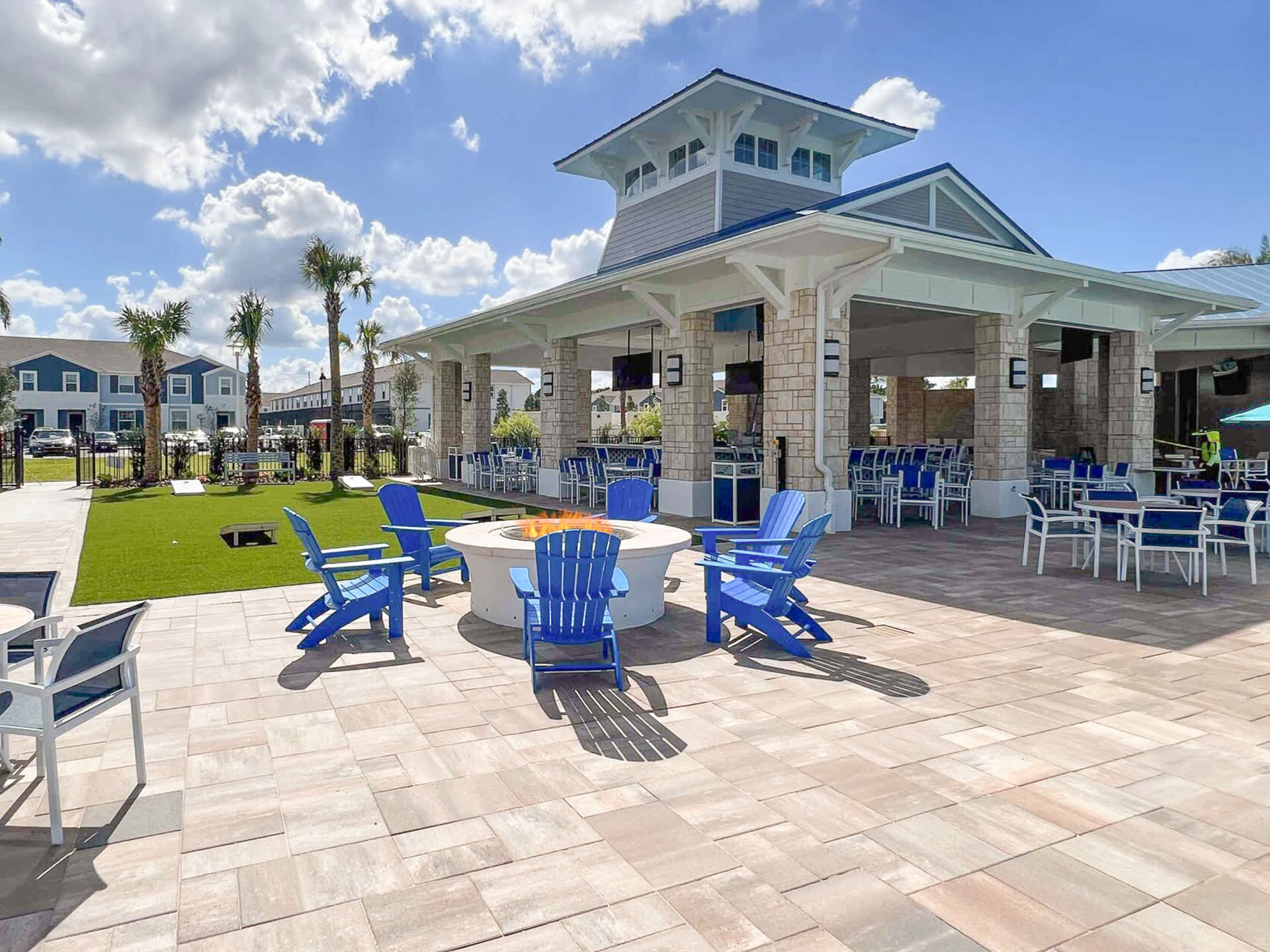 An inviting outdoor lounge area features a circular fire pit surrounded by blue Adirondack chairs on a paved patio, with a modern clubhouse building and palm trees under a partly cloudy sky.