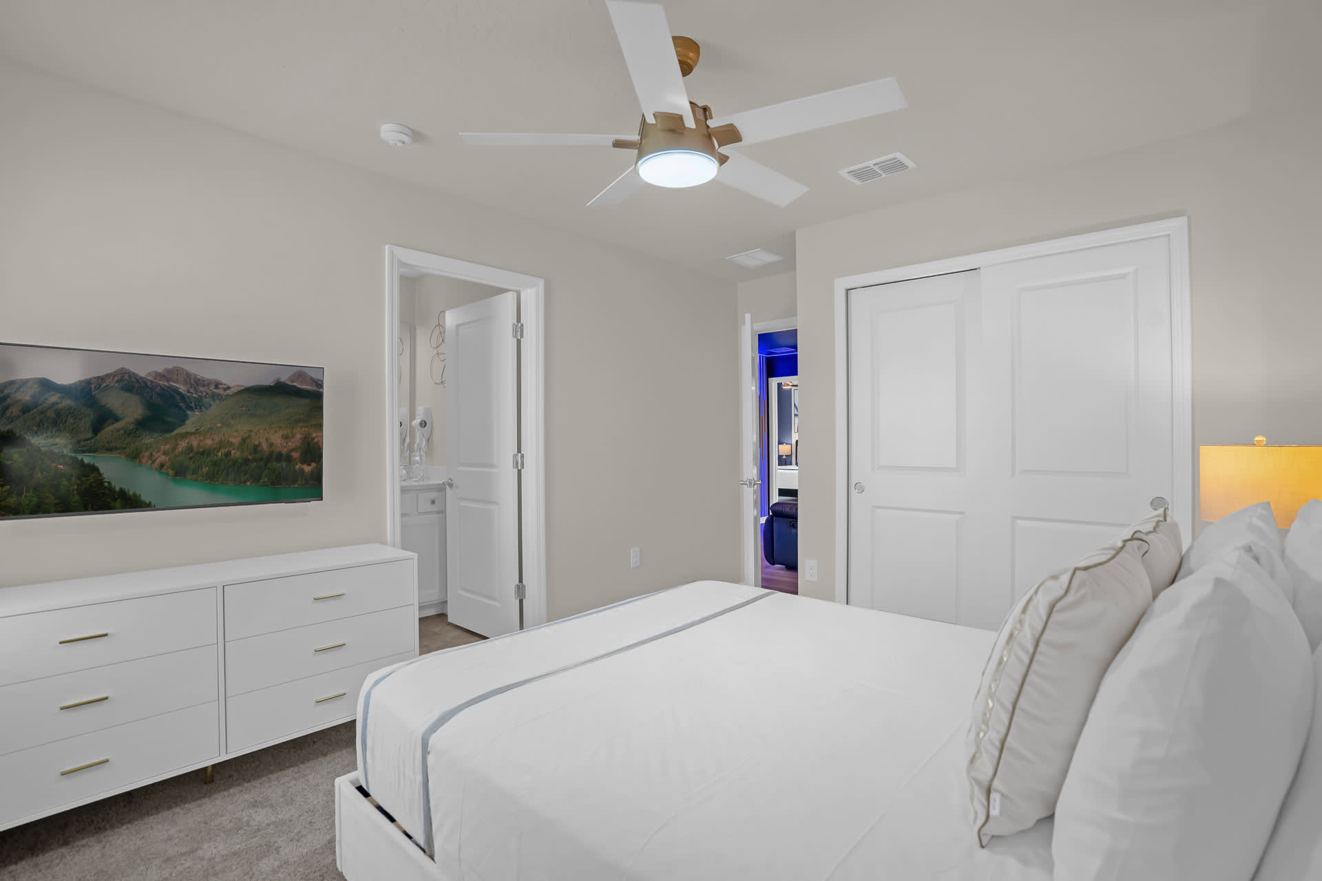 A bright bedroom with a large bed, a white dresser with a TV mounted above it, and a view into an adjacent bathroom, featuring a light-colored ceiling fan and carpeted floor.
