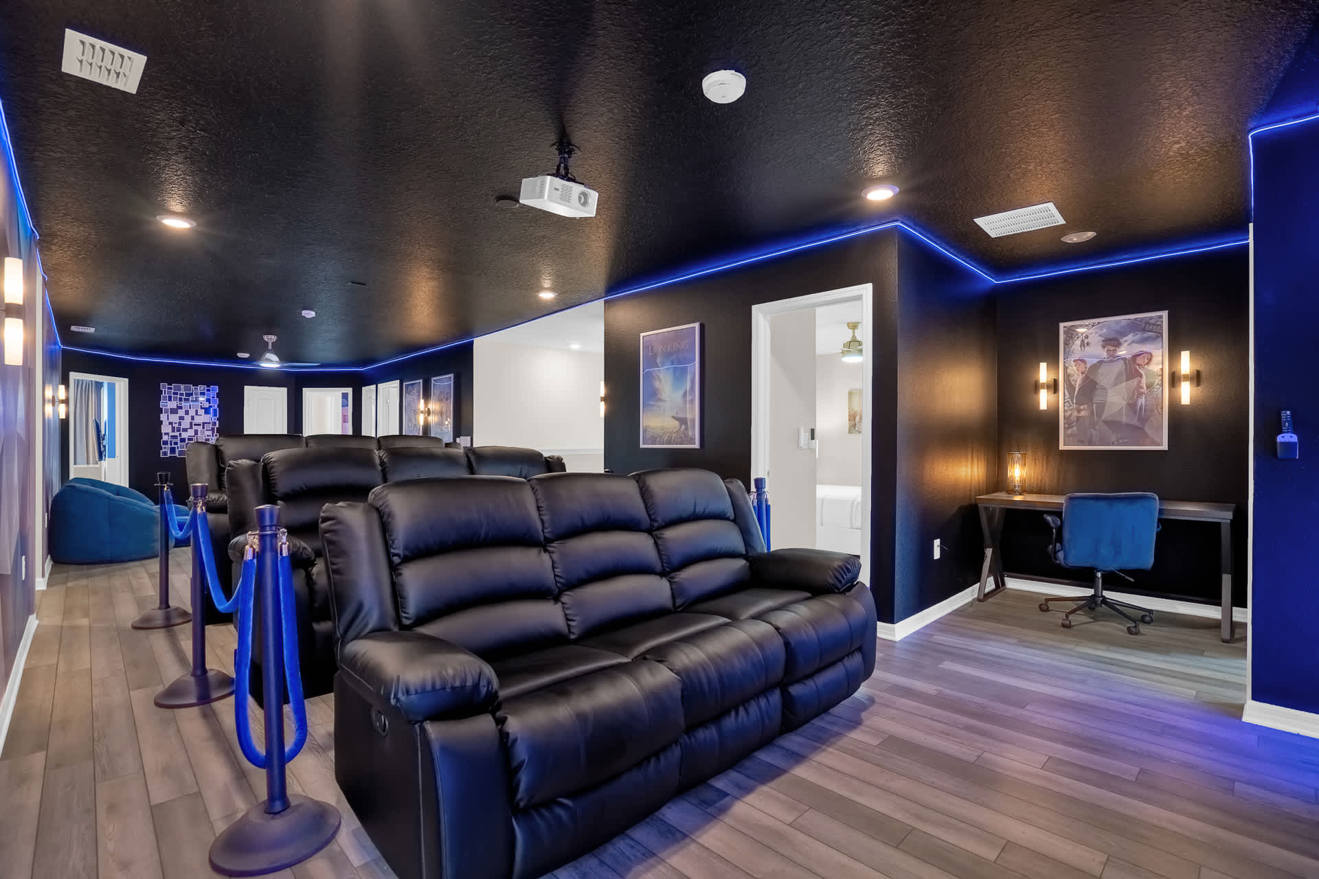 A home cinema room featuring multiple rows of black leather recliners, a projector mounted on the ceiling, and a small desk with a blue chair, all accented by blue LED strip lighting.
