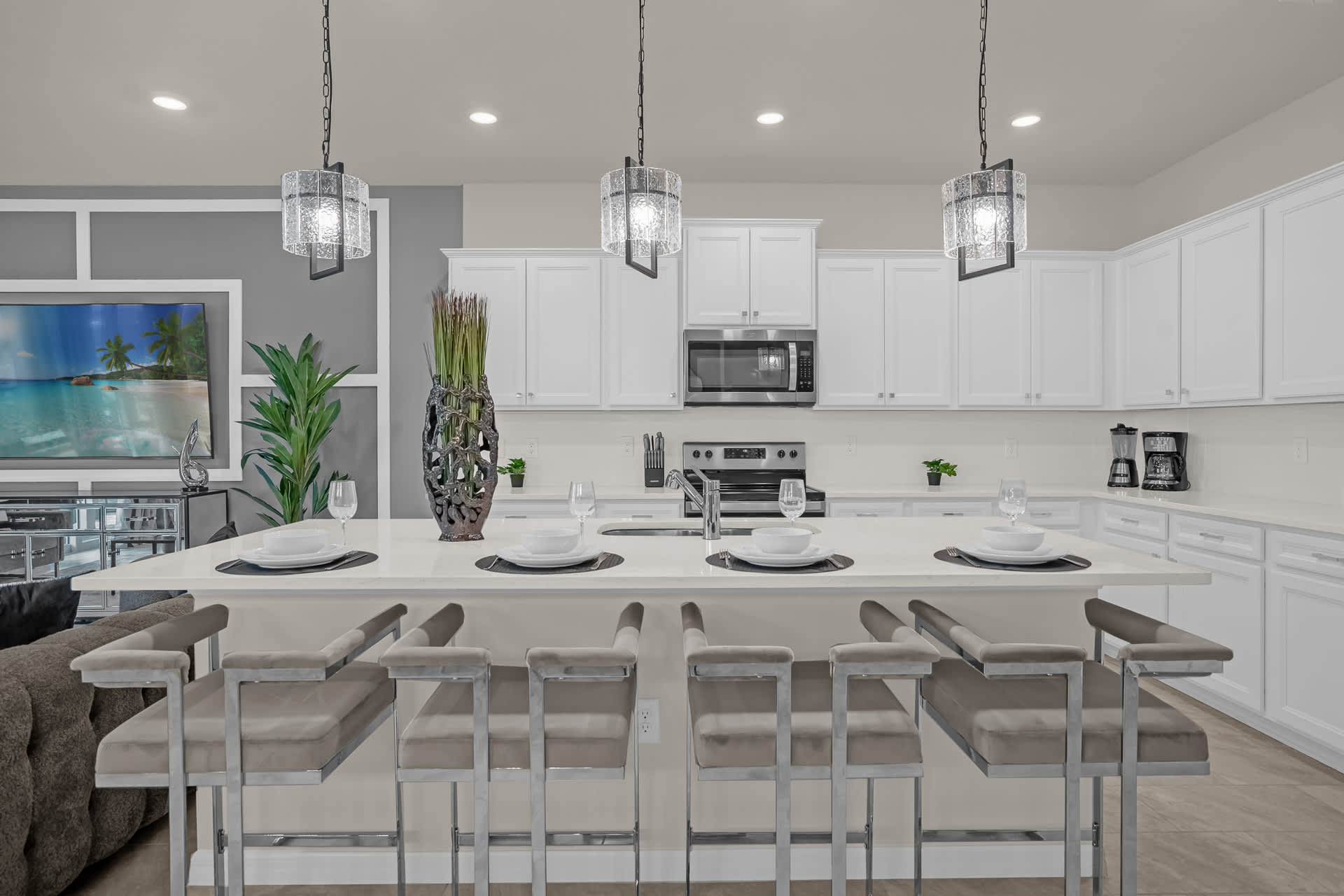A modern kitchen island with four gray bar stools, a sink, and pendant lighting, offering a view into the adjacent living room with a large TV.