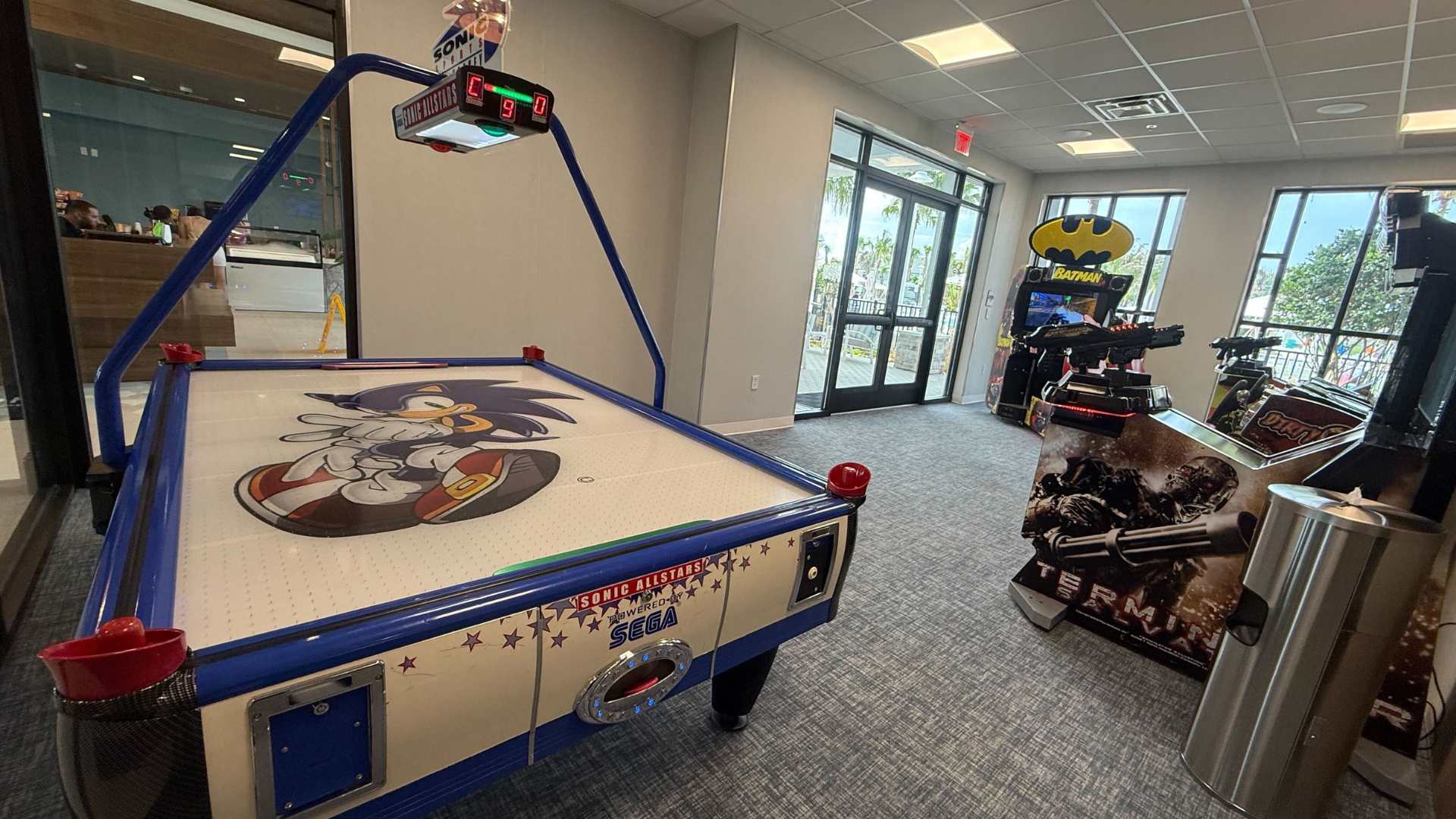 A "Sonic Allstars" air hockey table is prominently featured in this arcade room, with other arcade machines like "Terminator Salvation" and a Batman-themed game visible in the background.