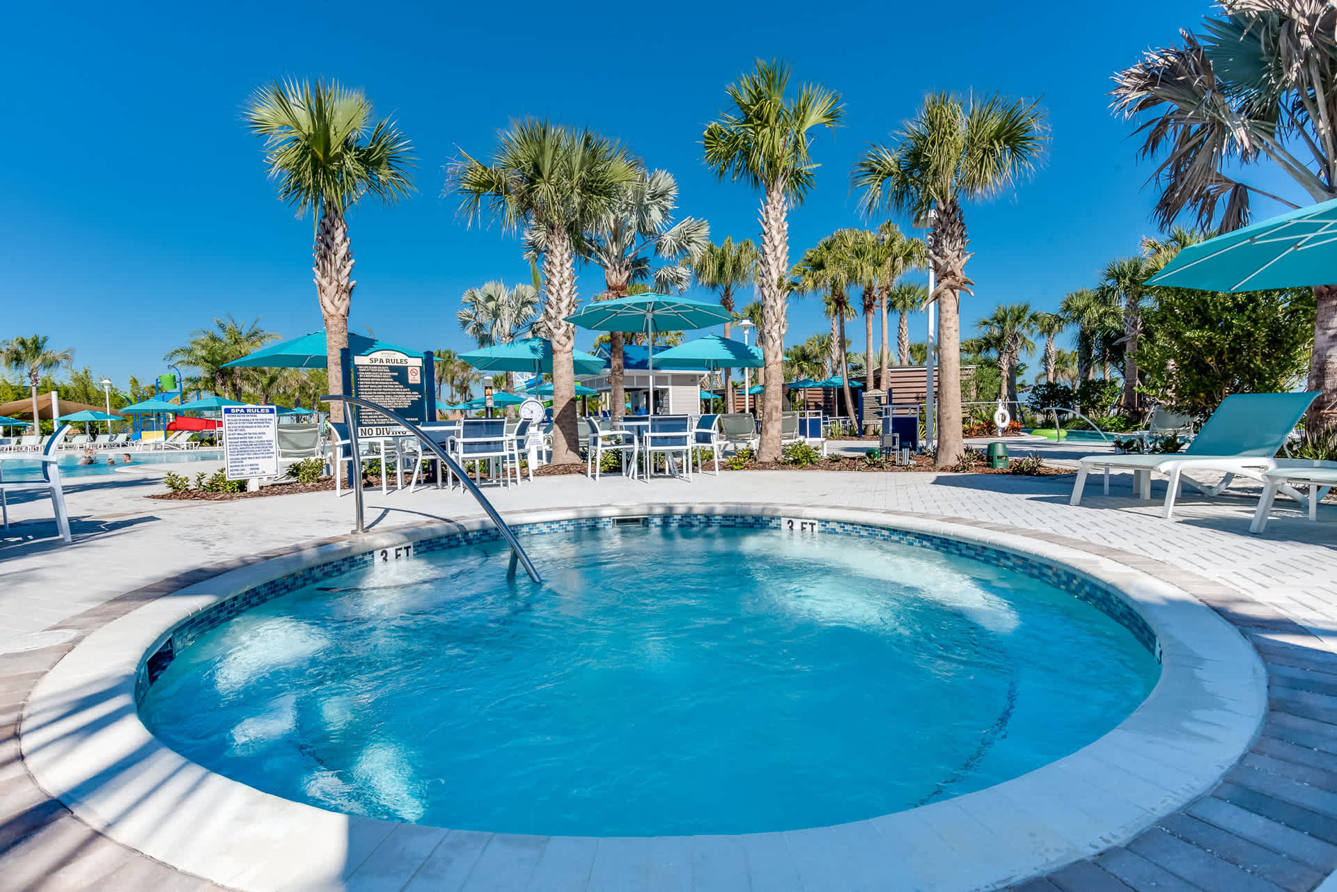 Circular outdoor hot tub with bubbling water, featuring stainless steel handrails, surrounded by light pavers, lounge chairs, and palm trees, with a "SPA RULES" sign visible in the background.