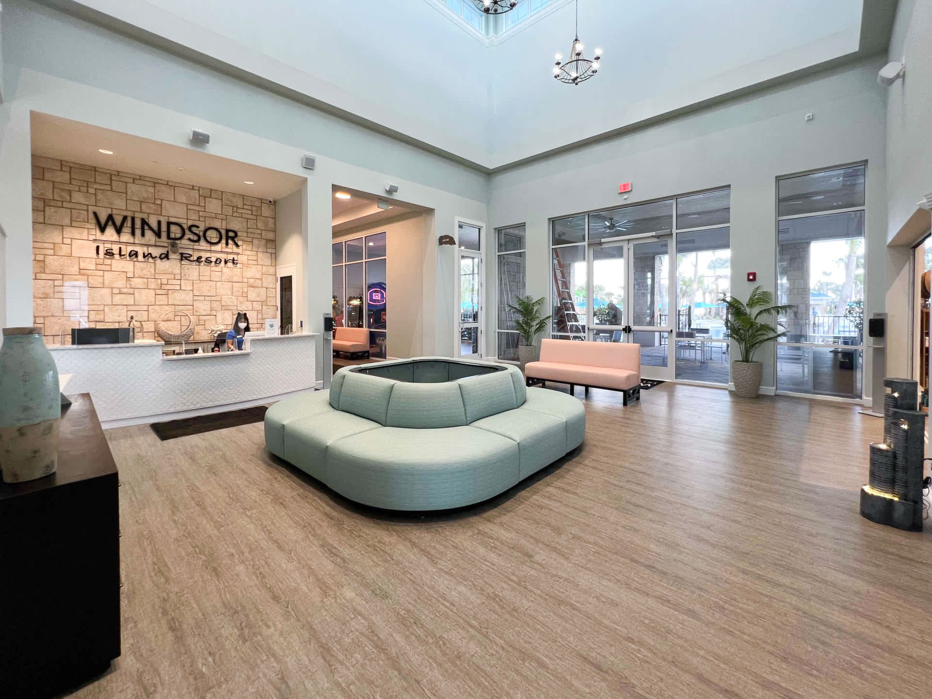 A bright and spacious resort lobby features a reception desk with a stone accent wall displaying "Windsor Island Resort", a large circular light-blue sofa, and glass doors leading to outdoor areas.