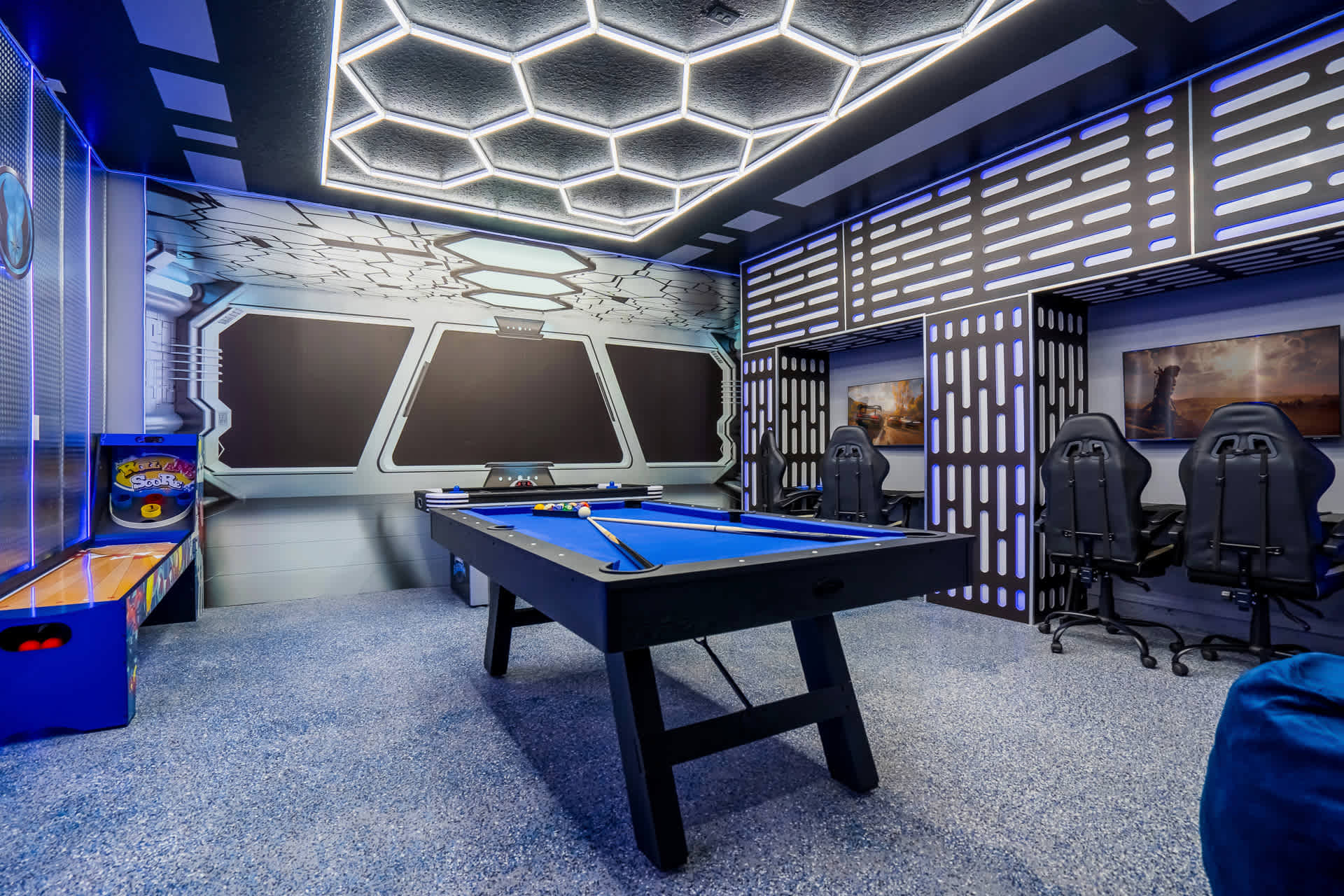 A sci-fi themed game room featuring a blue-felt pool table, a Skee-Ball arcade game, and two gaming stations with black chairs, all under a hexagonal LED ceiling light.