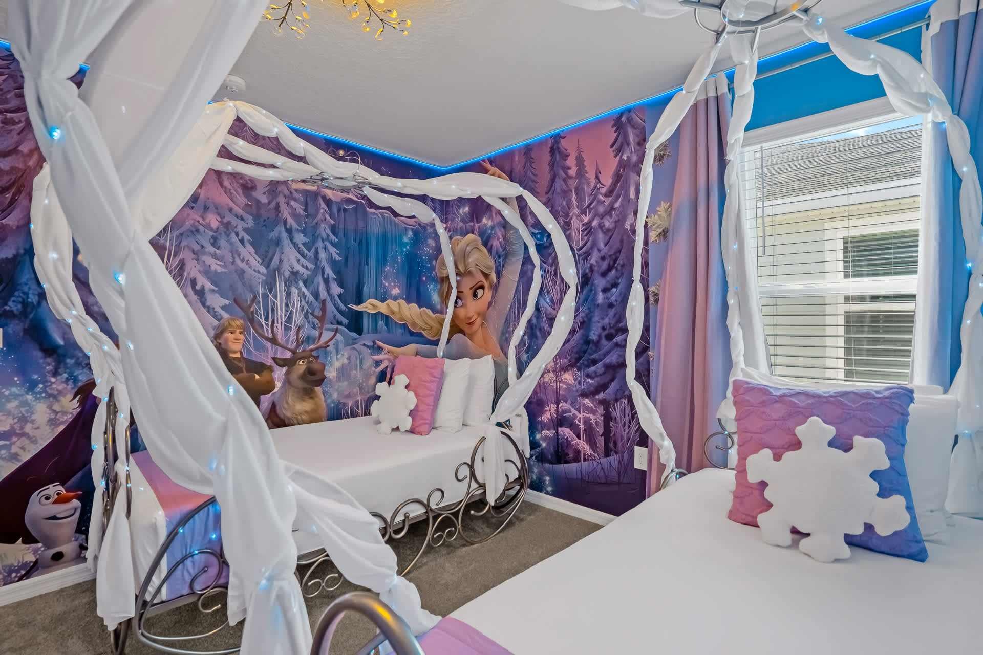 Detailed view of the 'Frozen' themed bedroom, showcasing two single beds with elegant canopy frames and a vibrant wall mural featuring Elsa, Anna, and Kristoff. The room includes a window with blinds and blue-themed decor.