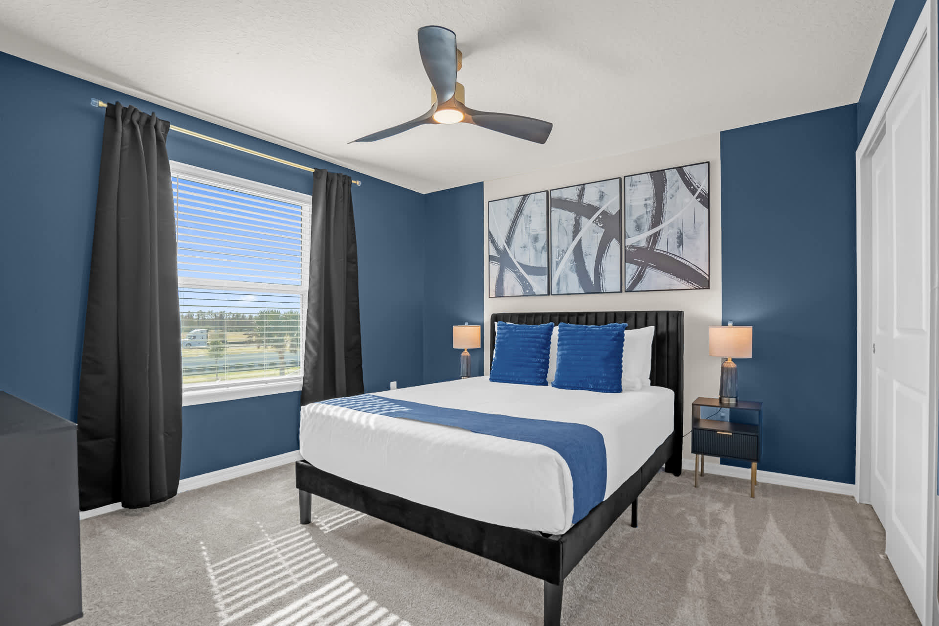 A stylish bedroom featuring a dark blue accent wall, a queen-sized bed with a black frame and blue pillows, two nightstands with lamps, and a large window with dark curtains.