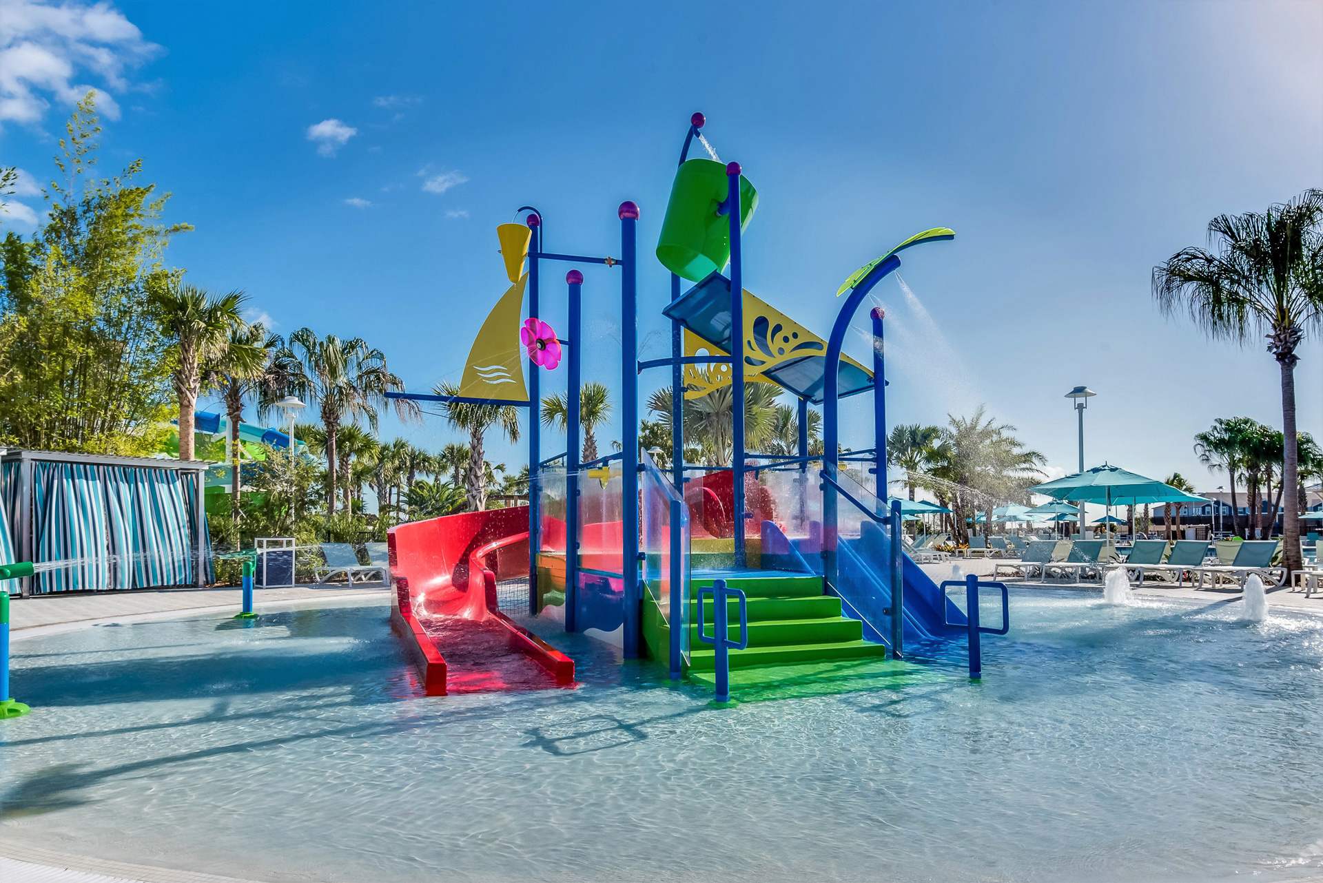 A vibrant outdoor splash pad features colorful water slides, climbing structures, and a large tipping bucket, set in a shallow pool under a bright blue sky with palm trees in the background.