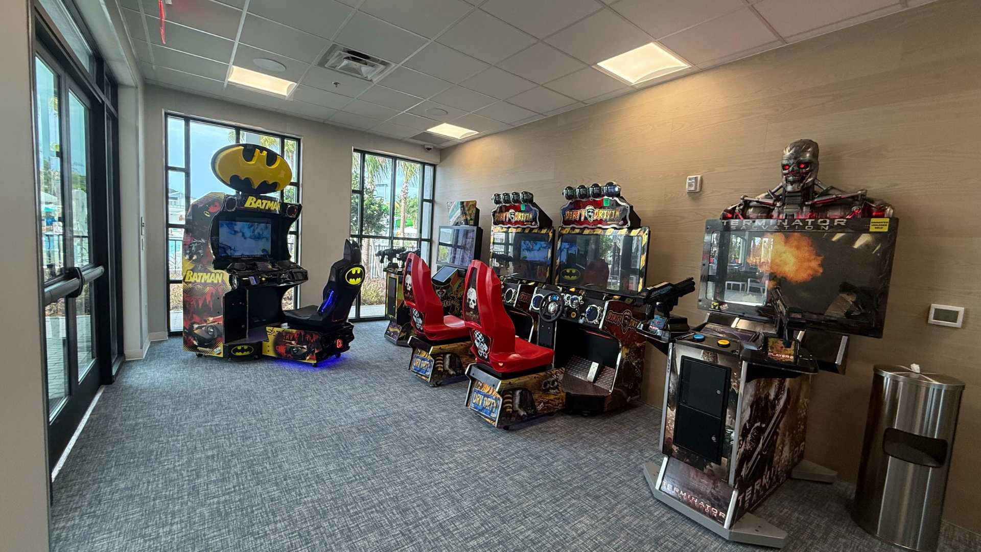 A well-lit game room features several arcade machines, including Batman, Dirty Driving, and Terminator, arranged on a gray patterned carpet. Large windows offer a glimpse of an outdoor pool area, and the walls are a mix of light wood paneling and white.
