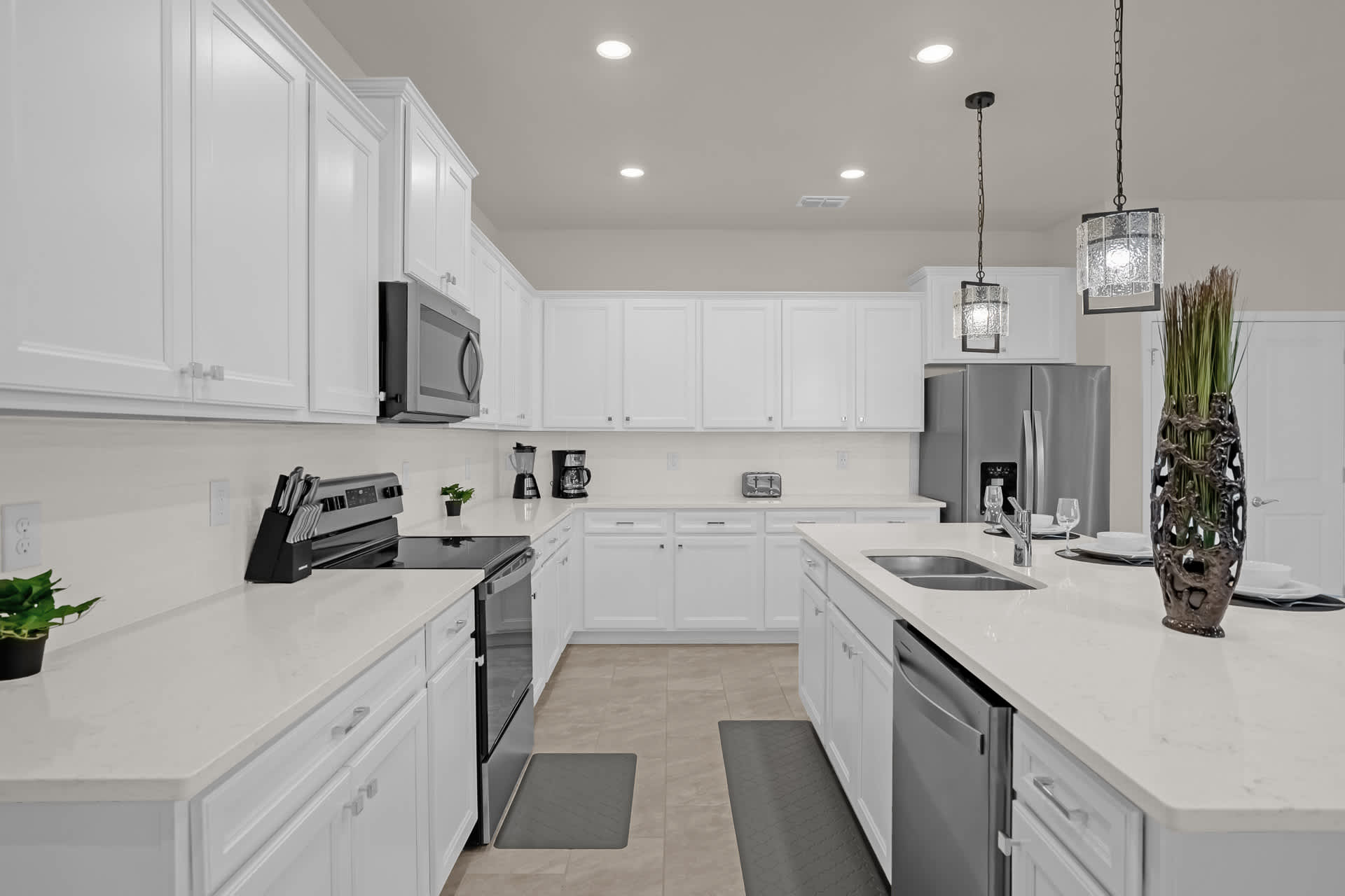A spacious, bright kitchen with white shaker cabinets, white countertops, stainless steel appliances including a refrigerator and oven, and a large island with a sink and pendant lights.