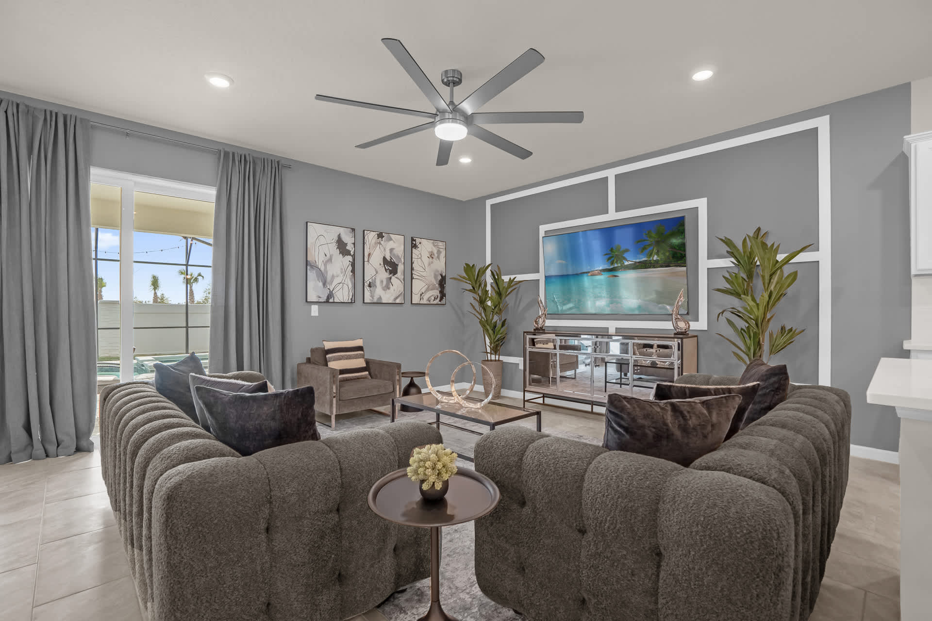 A contemporary living room featuring two plush gray sofas, a matching armchair, a large flat-screen TV mounted on a gray accent wall, and a modern ceiling fan, with large windows overlooking an outdoor area.