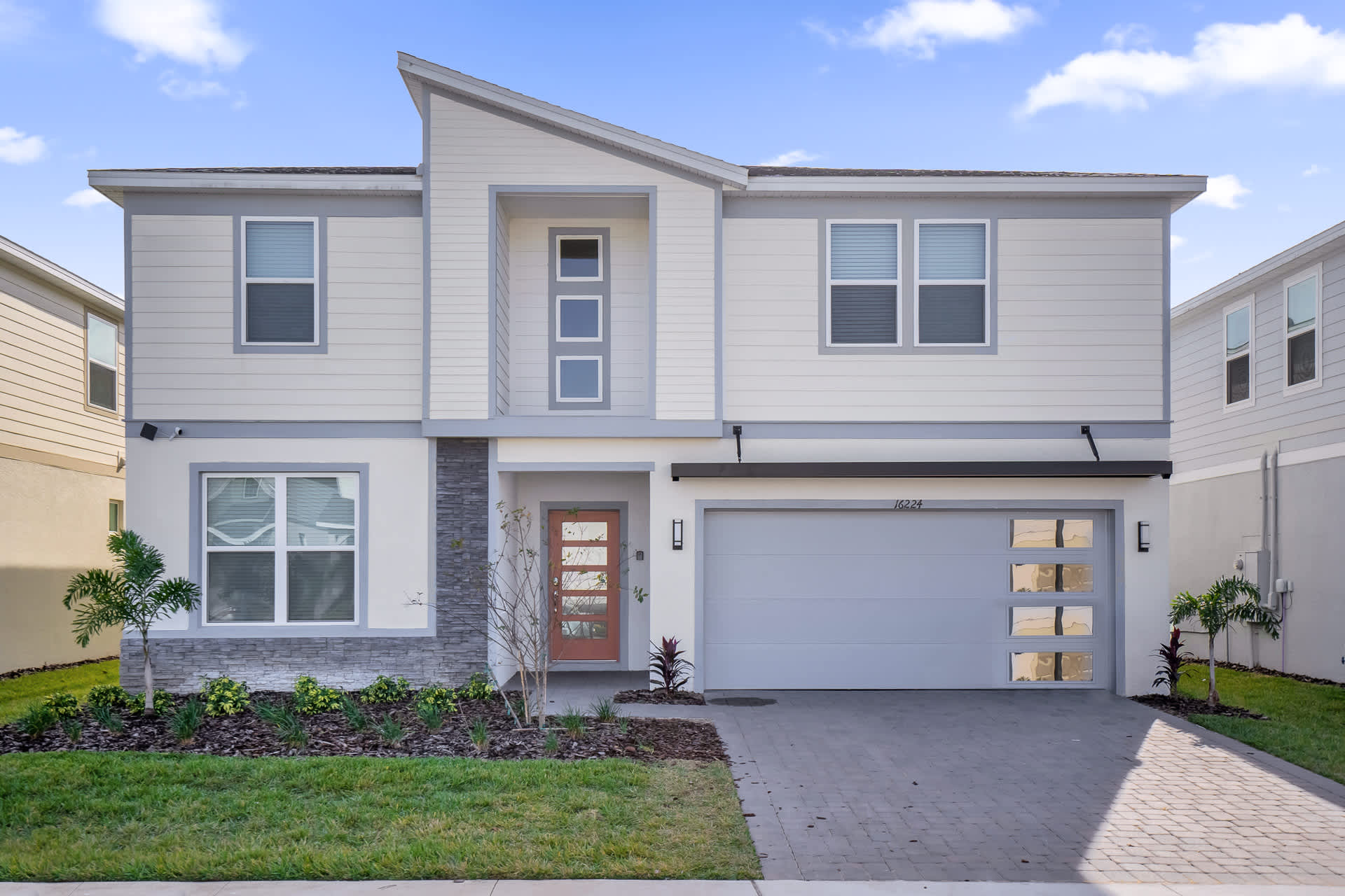 Modern two-story house exterior with light gray siding and white trim, featuring a prominent front entrance with a red door, a two-car garage, and a paved driveway.