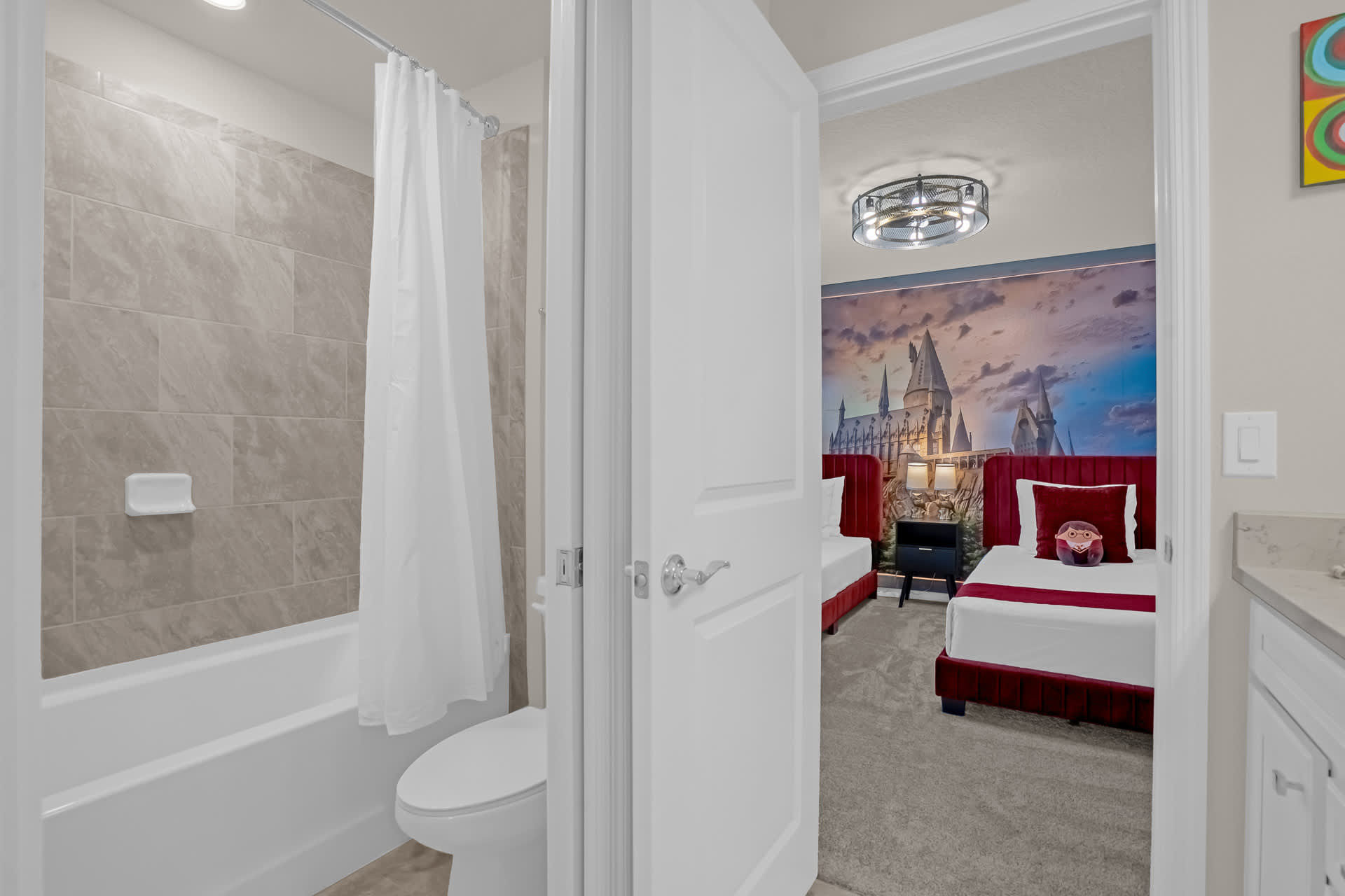 A modern bathroom featuring a tiled shower/bathtub combo with a white curtain and a white toilet, offering a glimpse into a themed bedroom with a large mural of a castle.