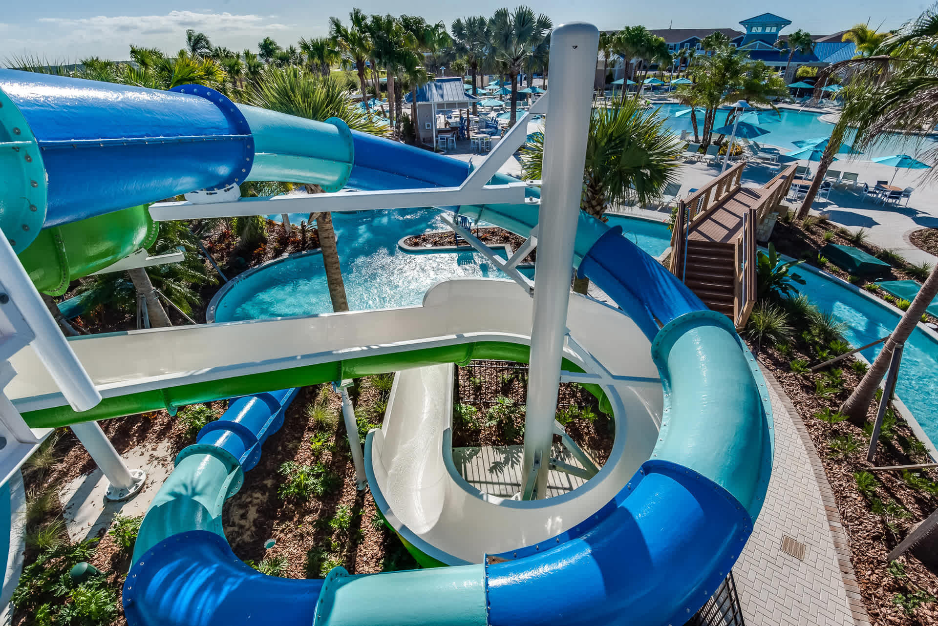 Dynamic view of a multi-colored water slide complex with blue, green, and white slides, offering a thrilling descent into a pool area, with a lazy river and lush landscaping visible below.