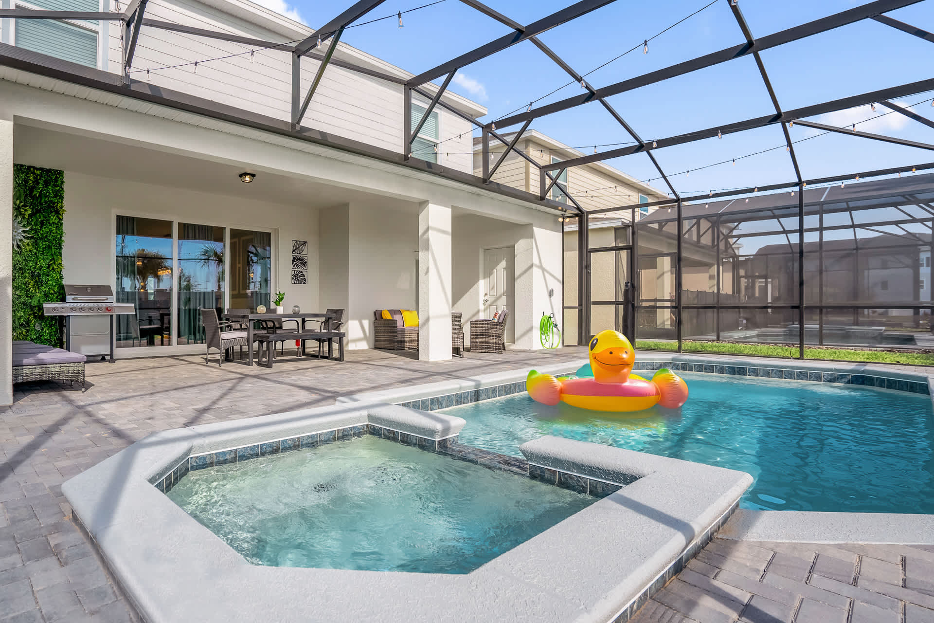 Spacious screened-in patio with a rectangular pool and integrated hot tub, featuring an outdoor dining set, lounge chairs, and a BBQ grill, all under a sunny sky.